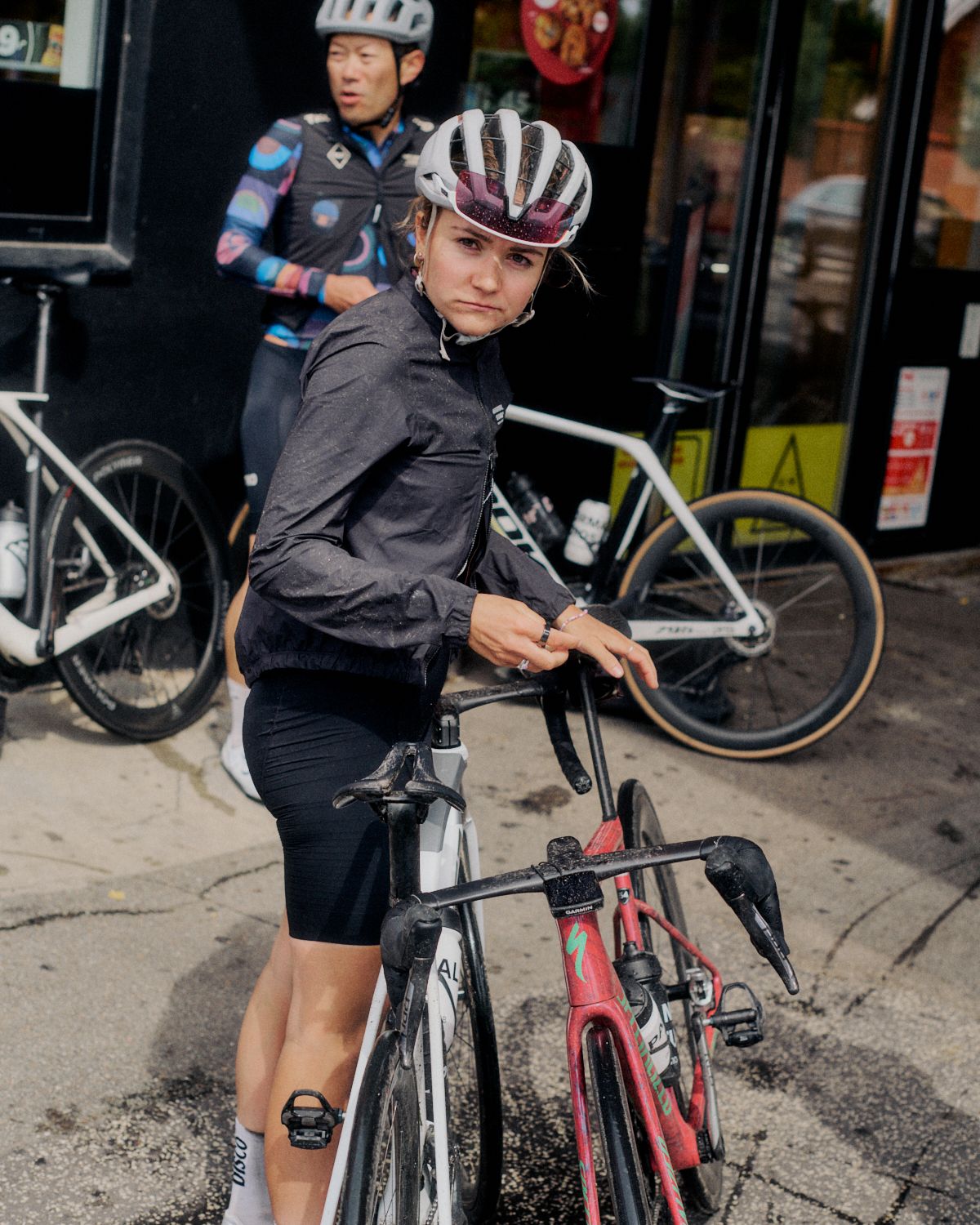 Model wearing dark grey cycling jacket with helmet, standing with bike in urban setting