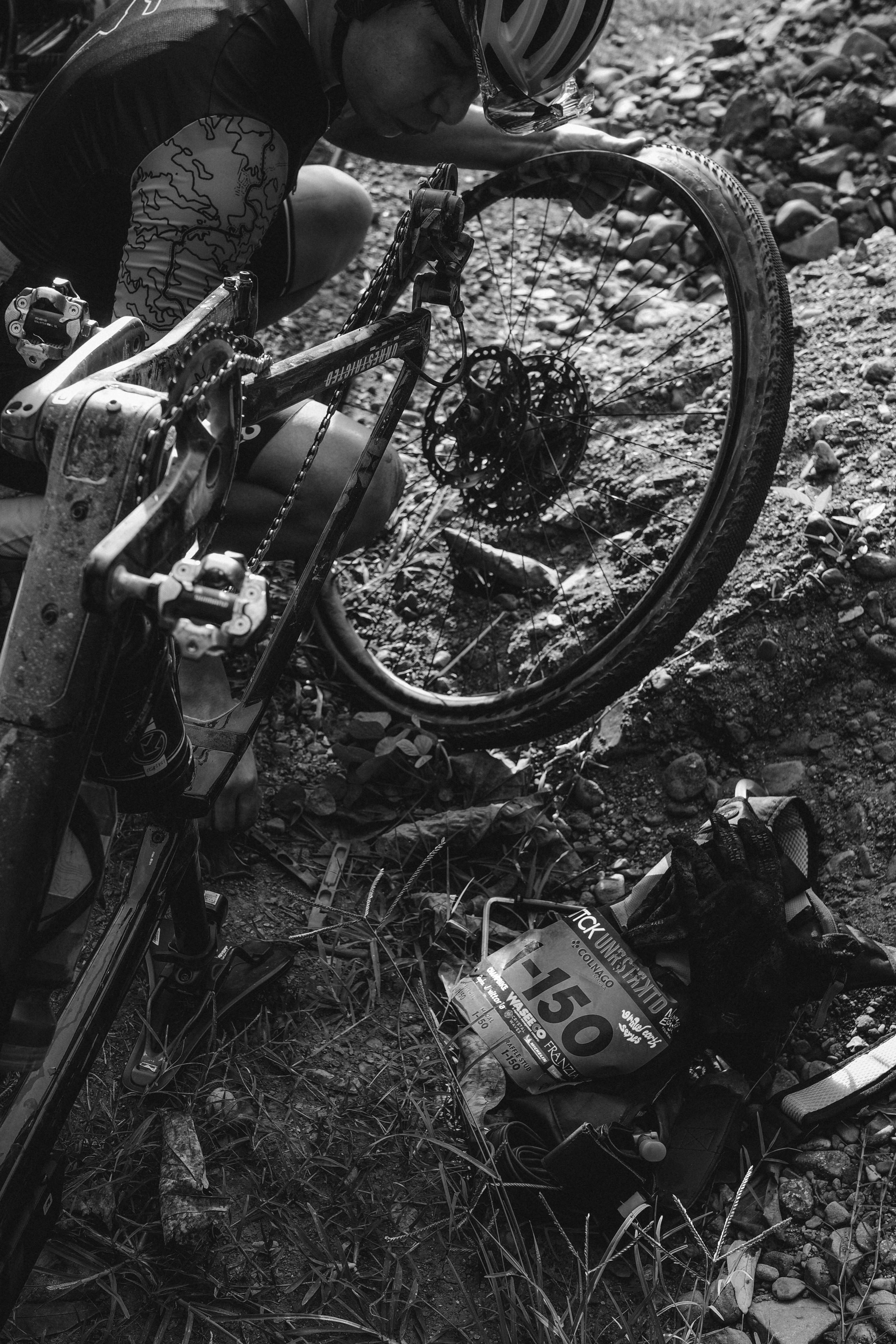 Cyclist with patterned leg warmers beside gravel bike on rough terrain in black and white