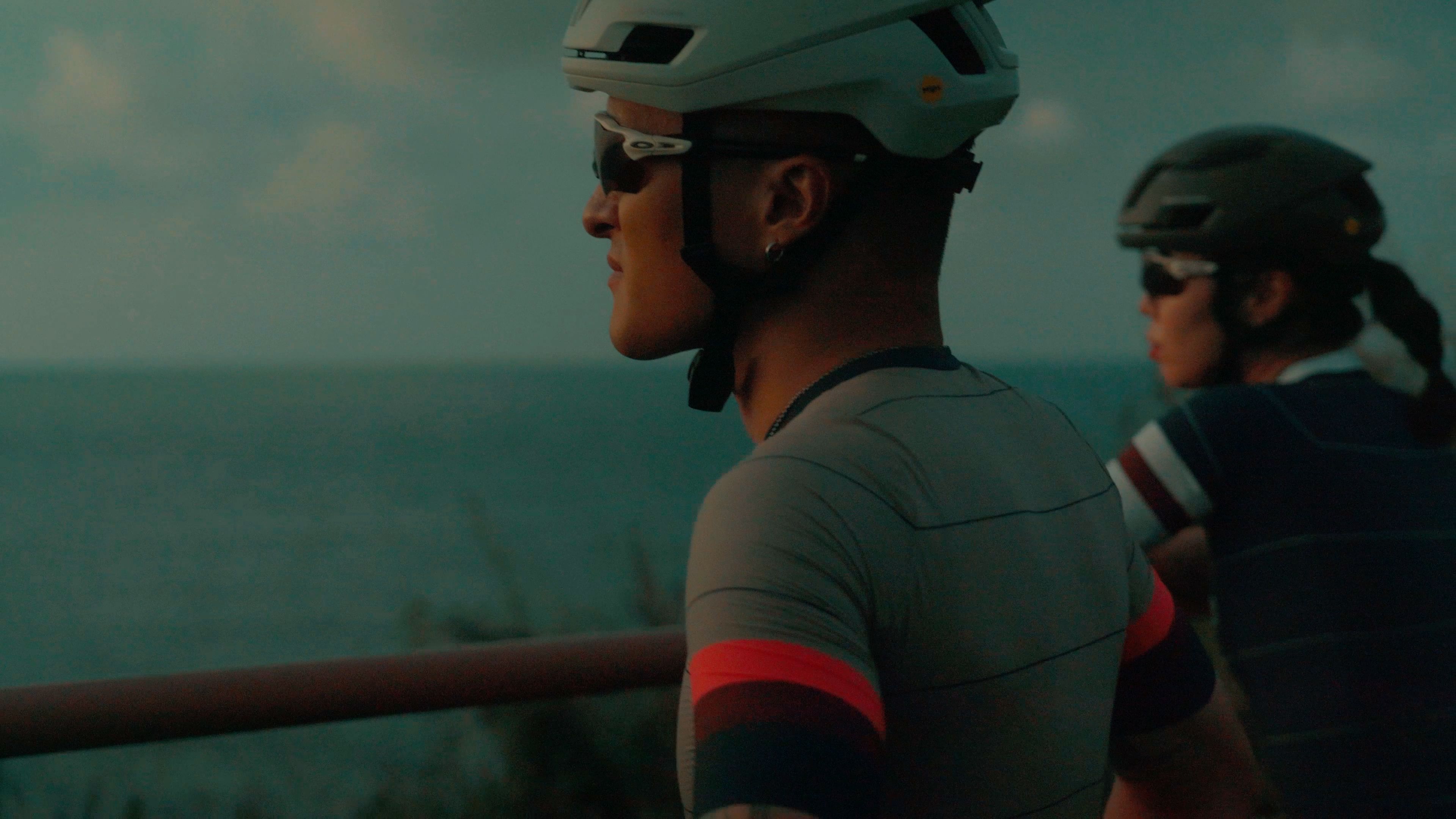 Models wearing grey cycling jerseys with red accents and helmets against teal sky backdrop