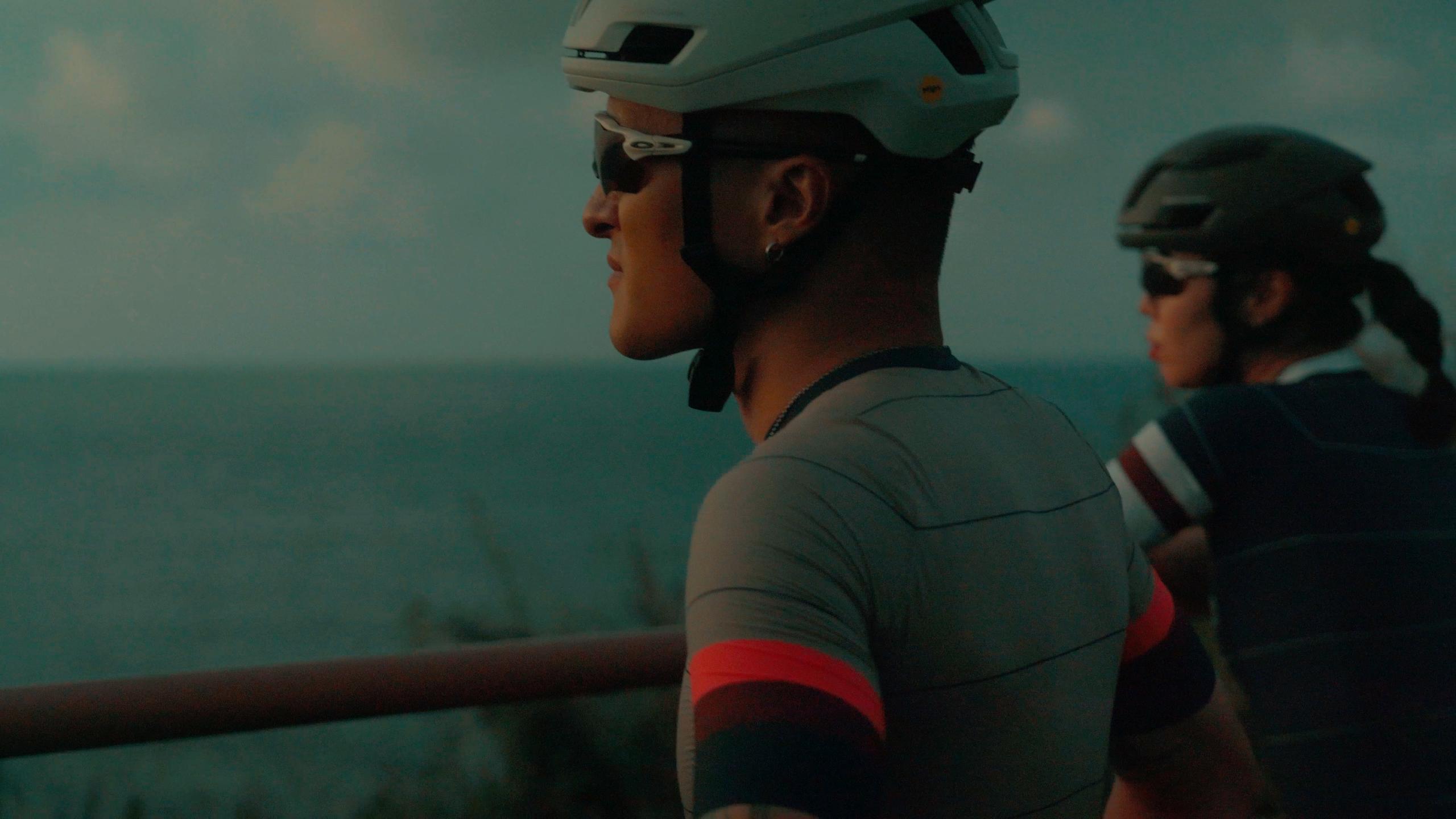 Models wearing grey cycling jerseys with red accents and helmets against teal sky backdrop
