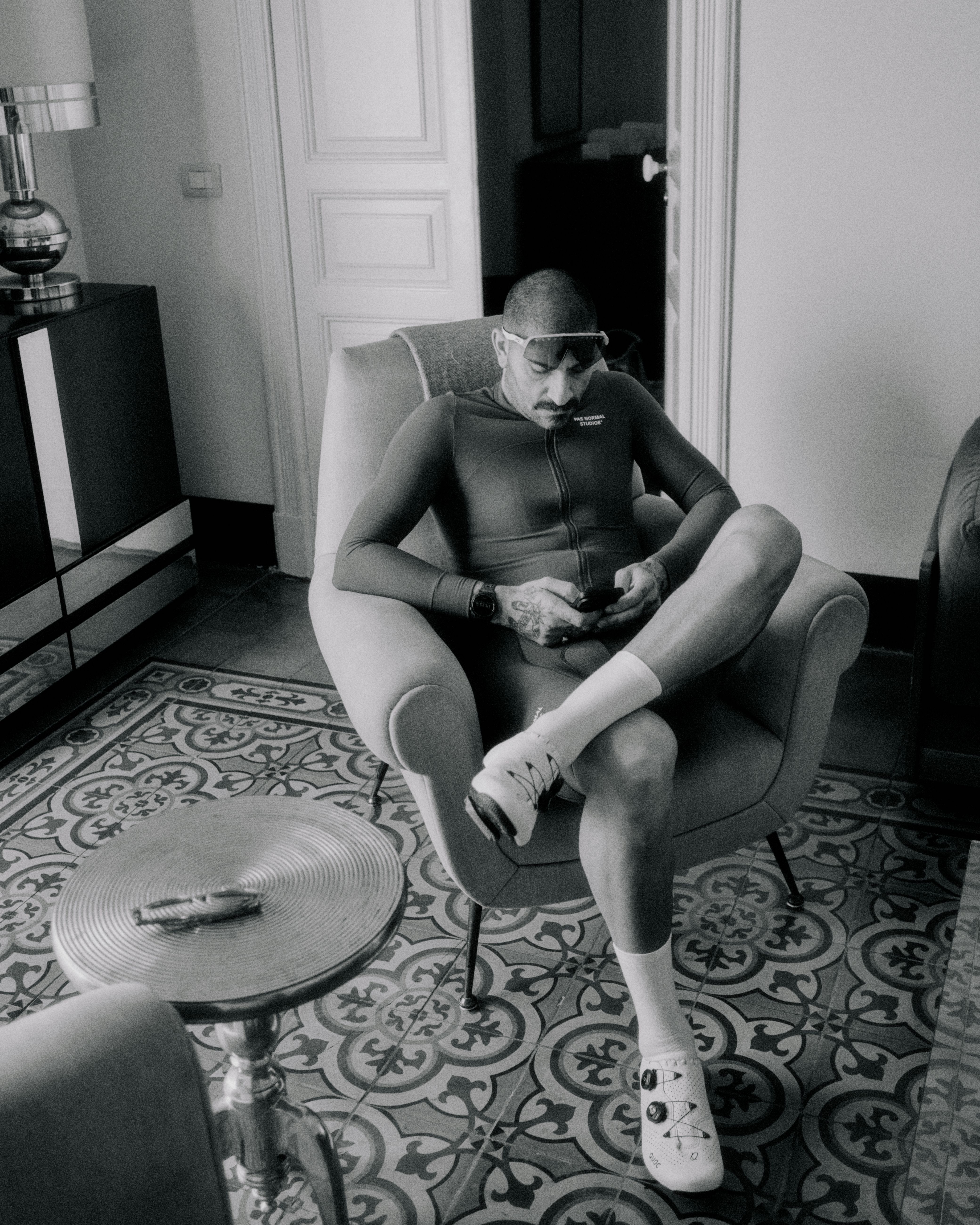 Model wearing dark cycling jersey and shorts, seated in chair with white cycling shoes in black and white photo