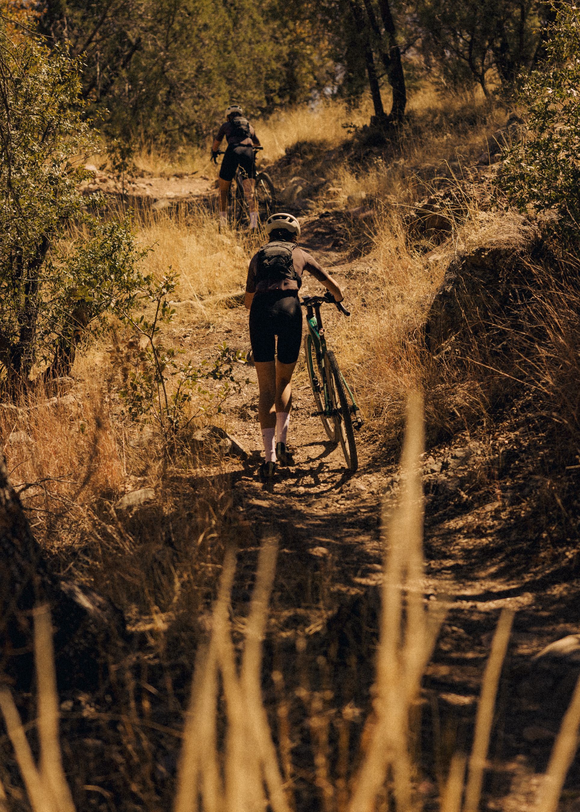 Model wearing dark cycling kit walking bike up rocky trail in golden mountain terrain