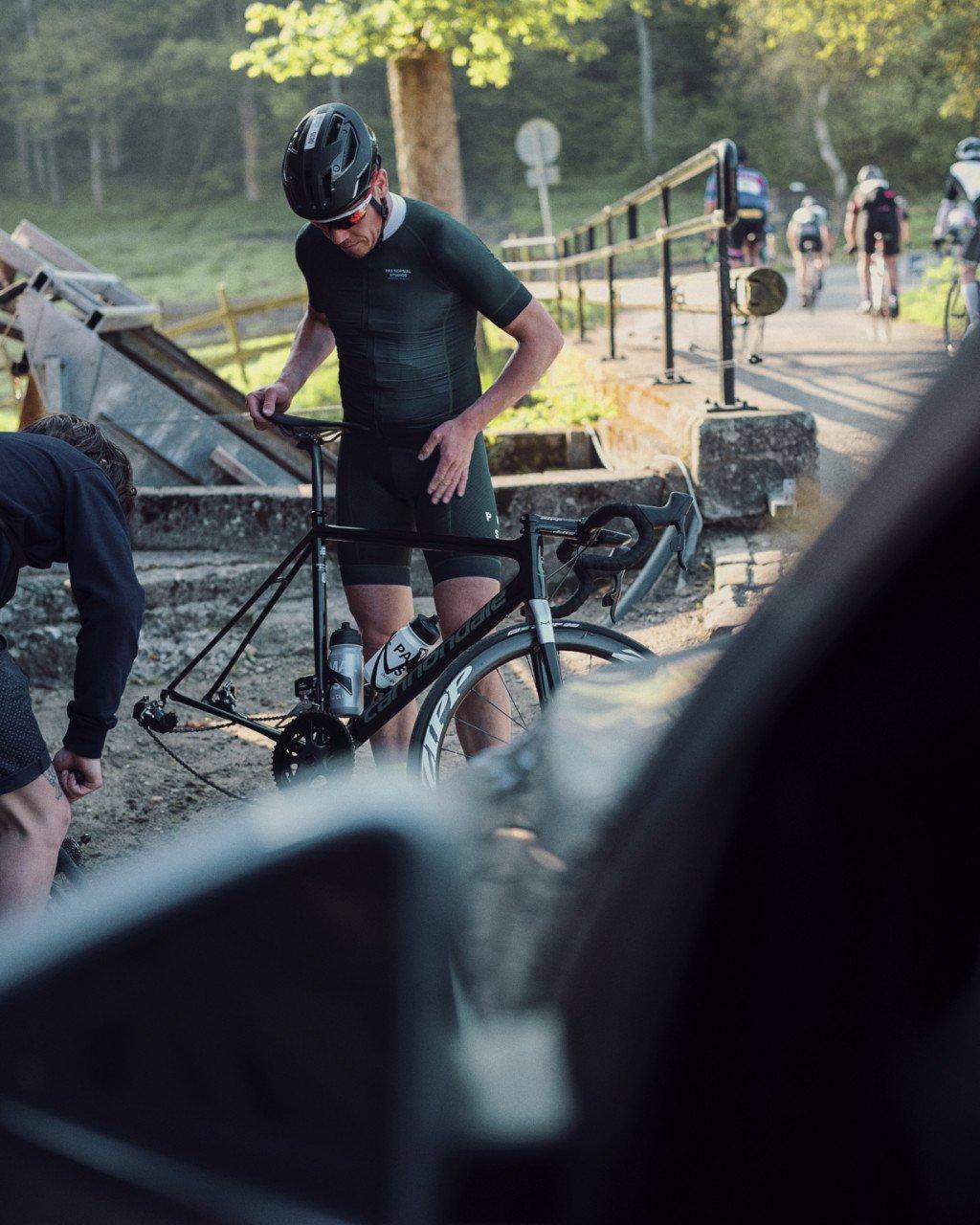 Model wearing green short sleeve jersey preparing time trial bike at rural bridge with other cyclists in background