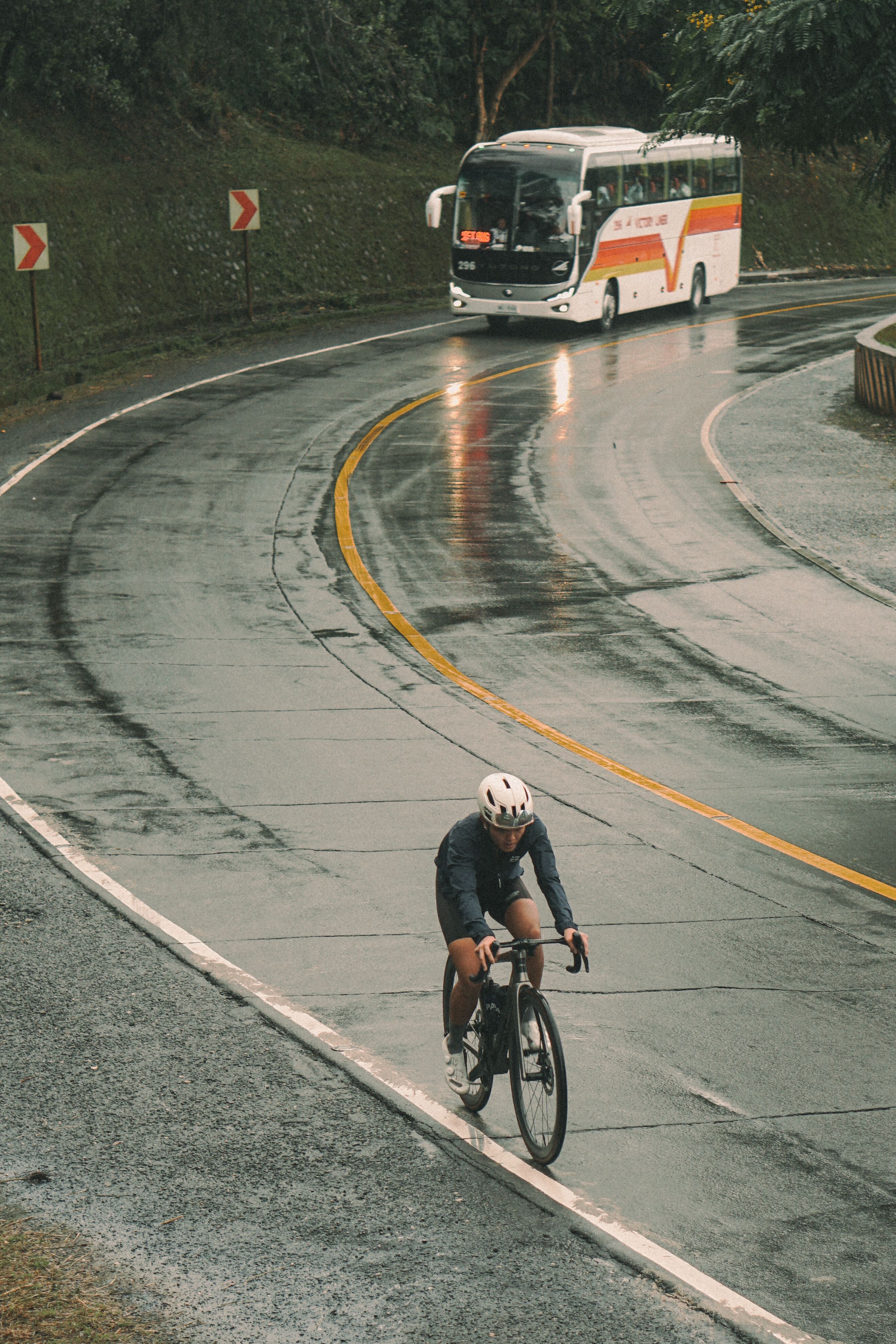 Cyclist in dark apparel riding on wet mountain road with bus in background