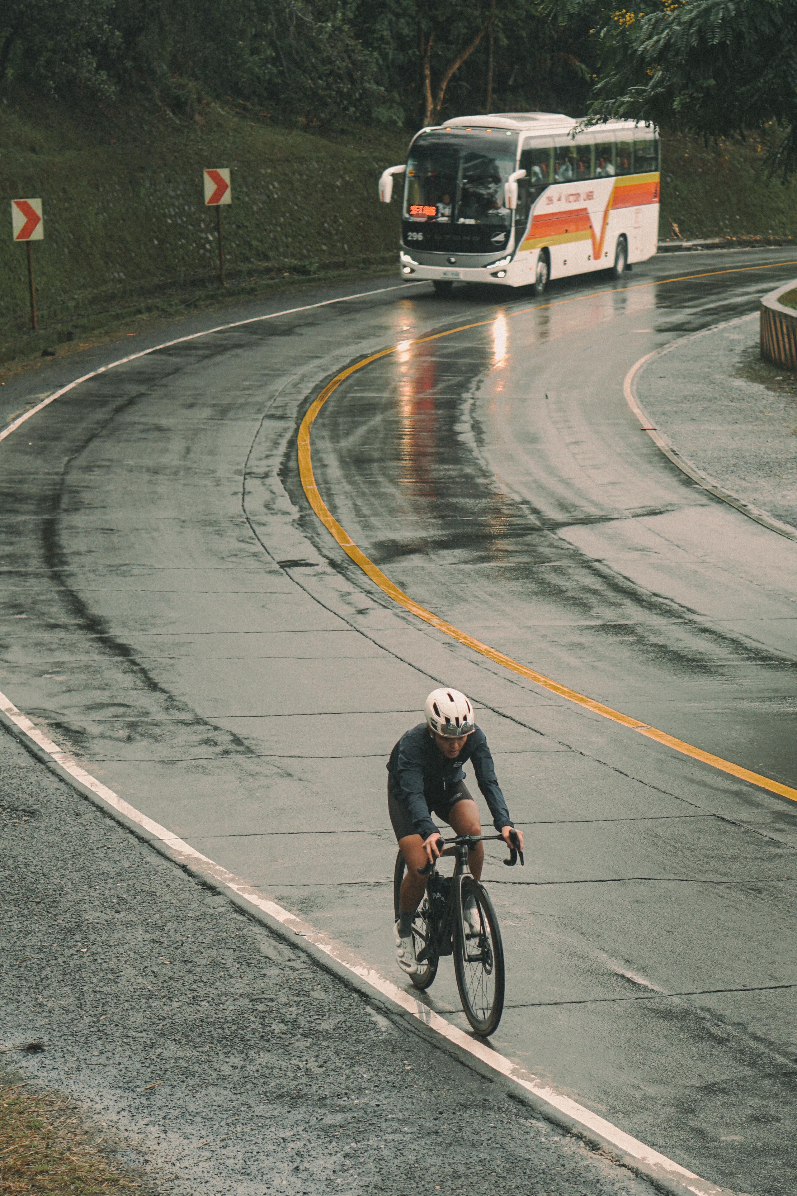 Cyclist in dark apparel riding on wet mountain road with bus in background