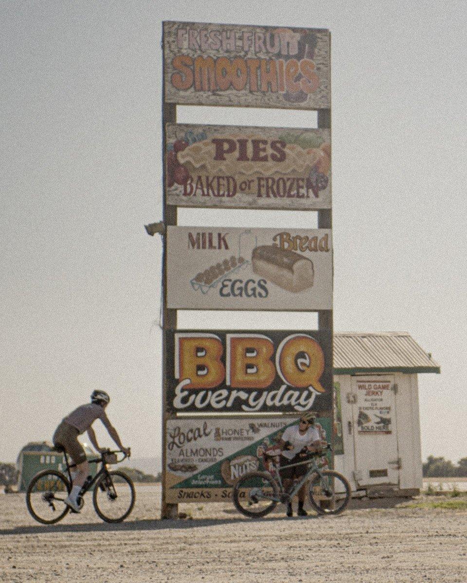 Cyclists at roadside diner sign advertising smoothies, pies, milk, eggs and BBQ
