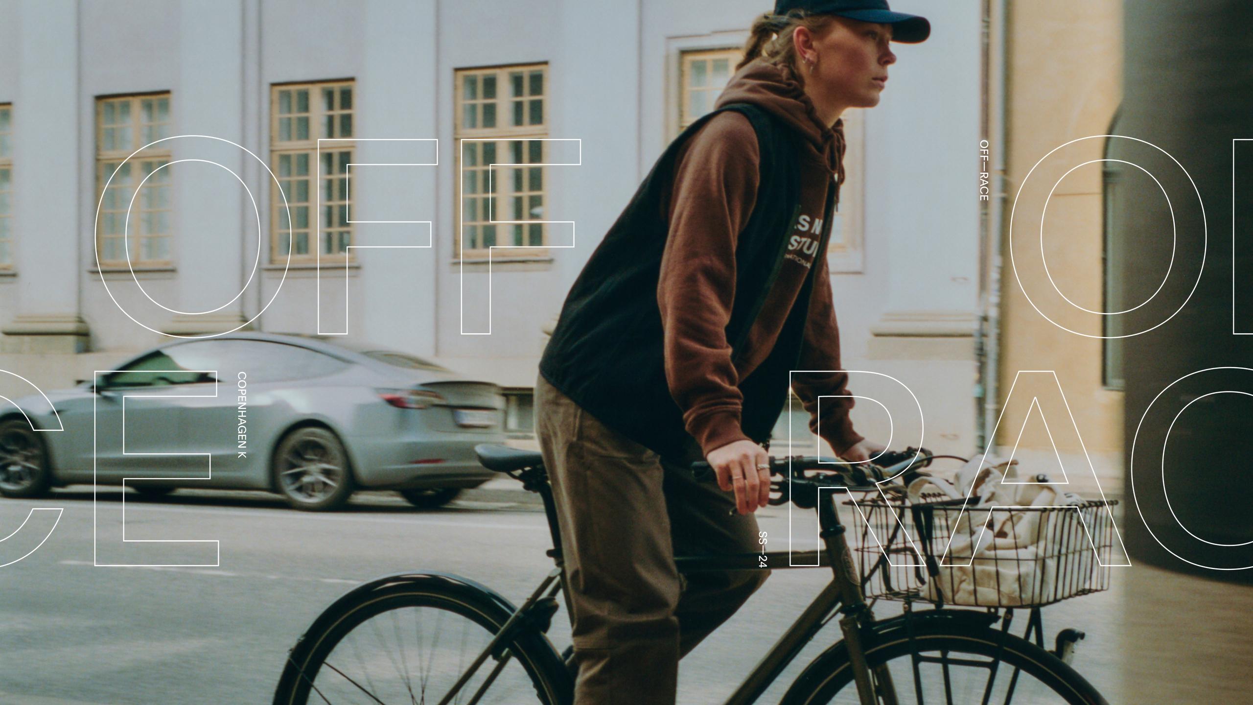 Model wearing brown hoodie and black cap with bike on Copenhagen street
