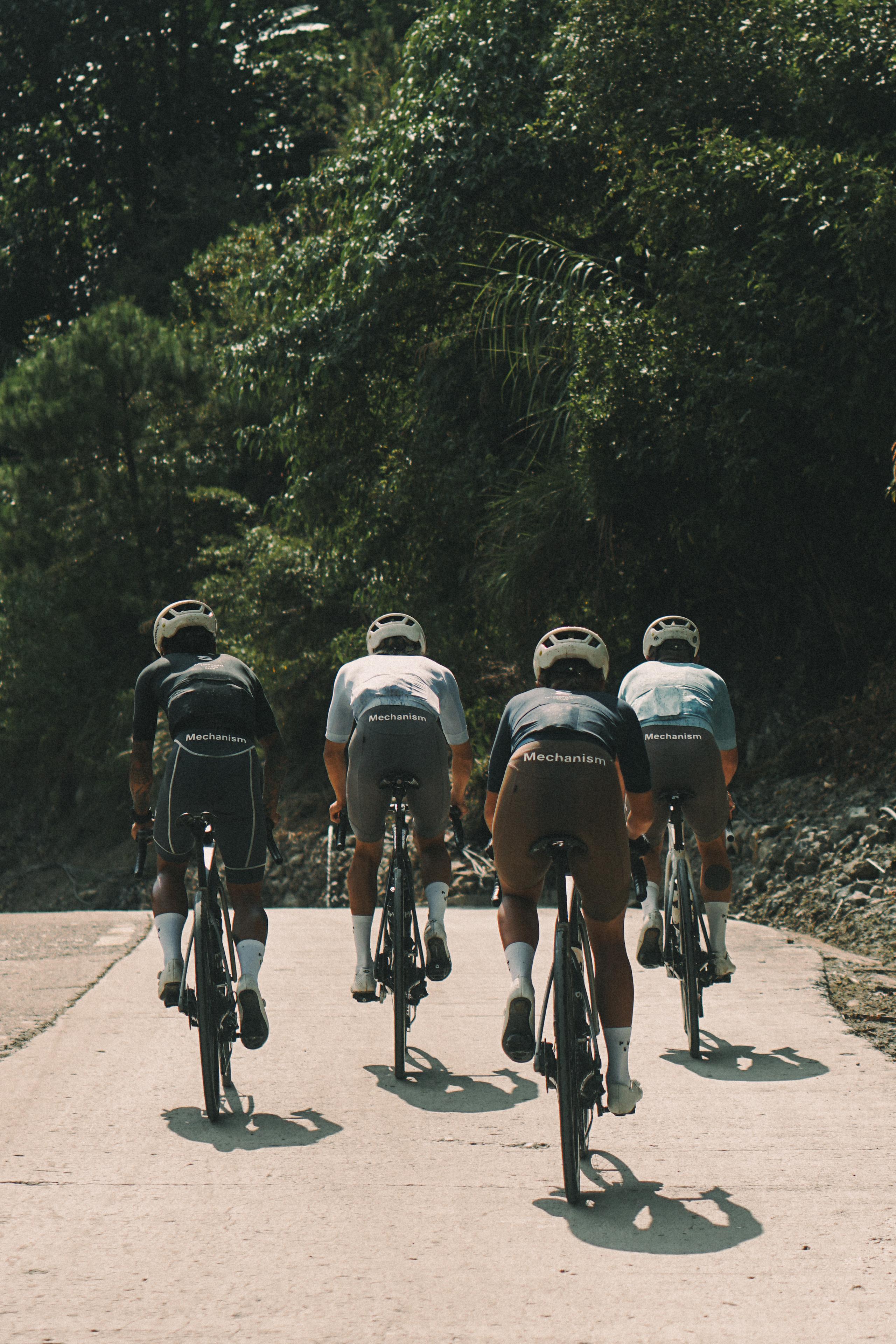 Four cyclists wearing Mechanism jerseys cycling on road through lush green landscape