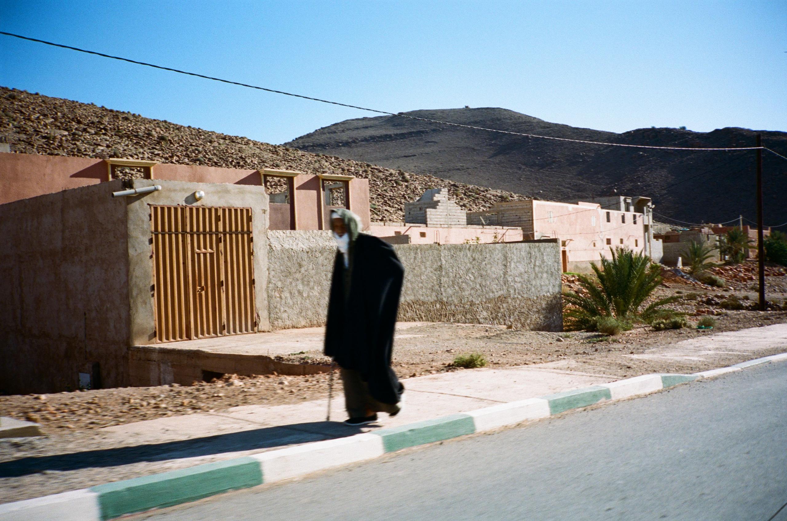 Model wearing black cycling attire walking on desert road with traditional buildings and mountains in background