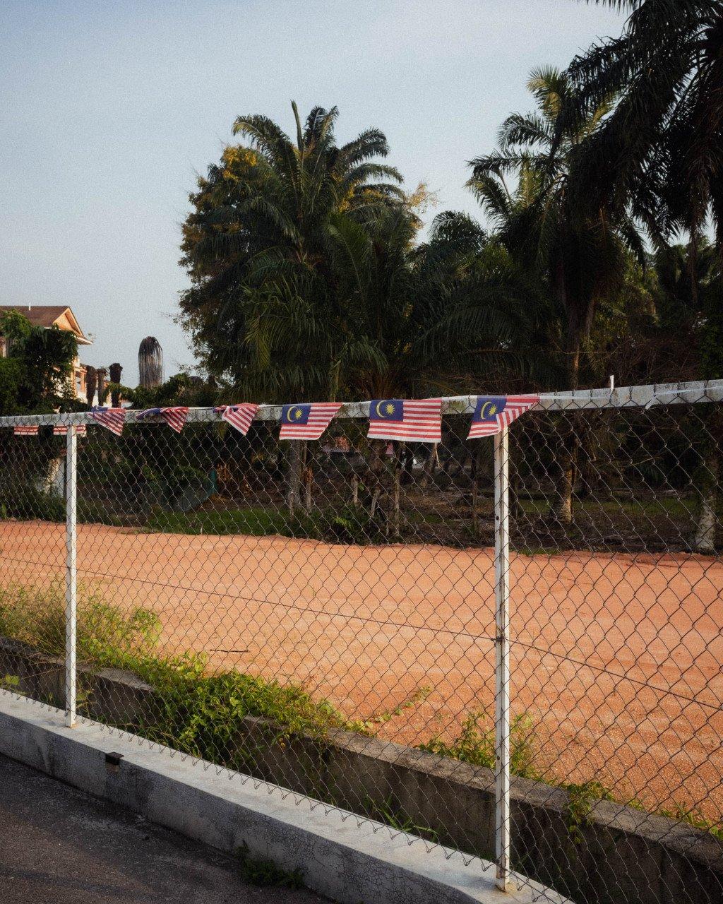 Tennis court with Malaysian flags hung on chain-link fence, palm trees in tropical setting