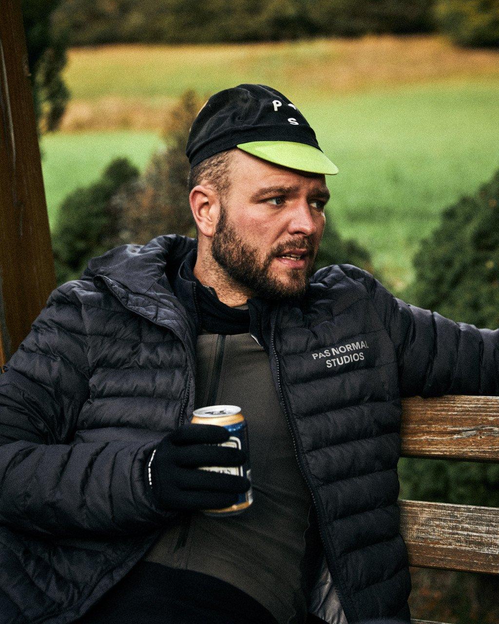Model wearing black quilted down jacket and cap with lettering, sitting at wooden table outdoors
