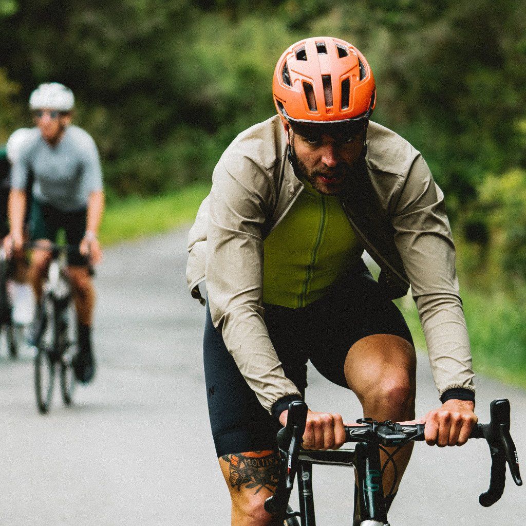 Model wearing tan long-sleeve cycling jersey with black bib shorts and orange helmet riding on road
