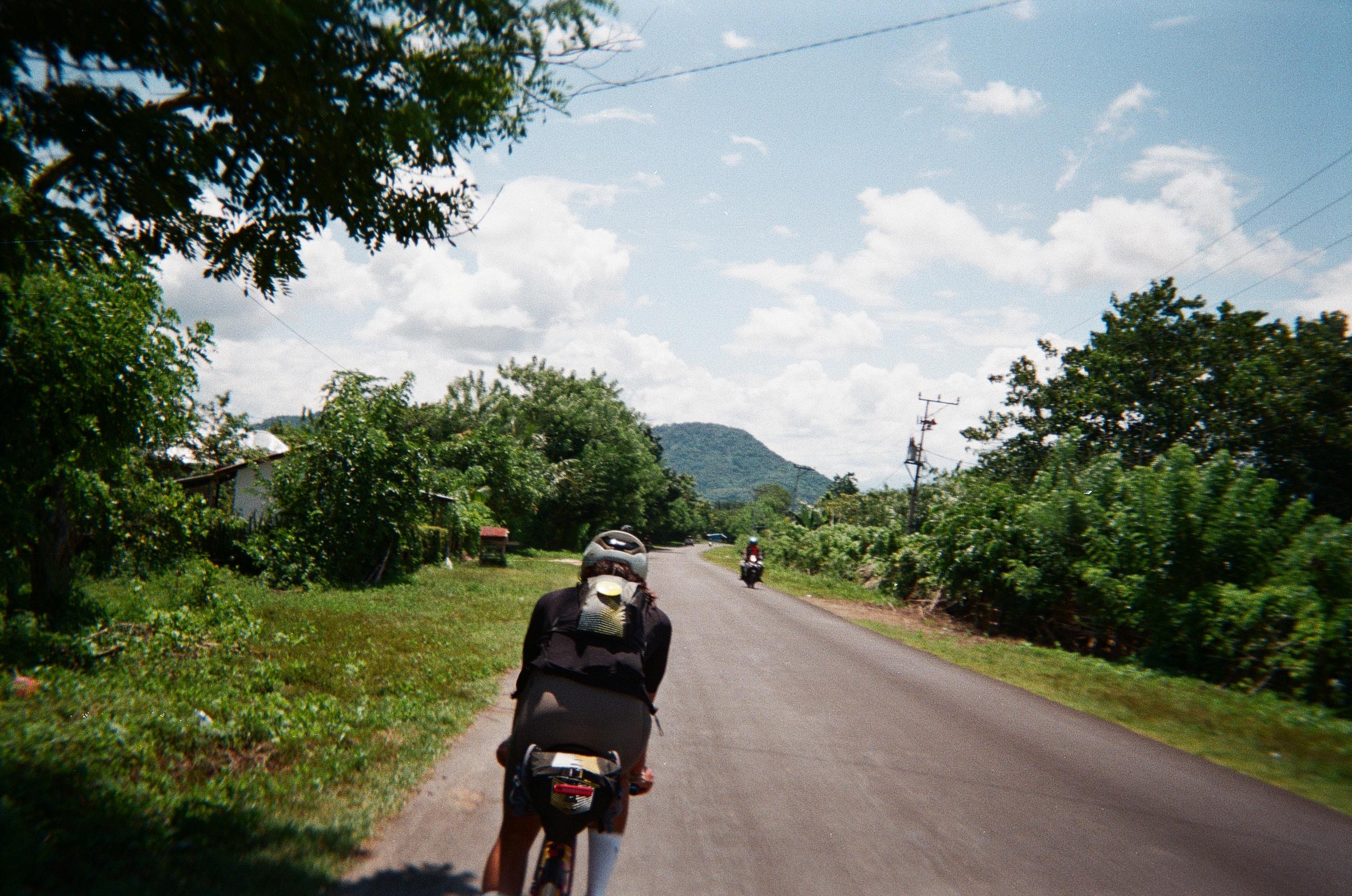 Cyclist in dark kit riding on rural road through lush green landscape with hills in background