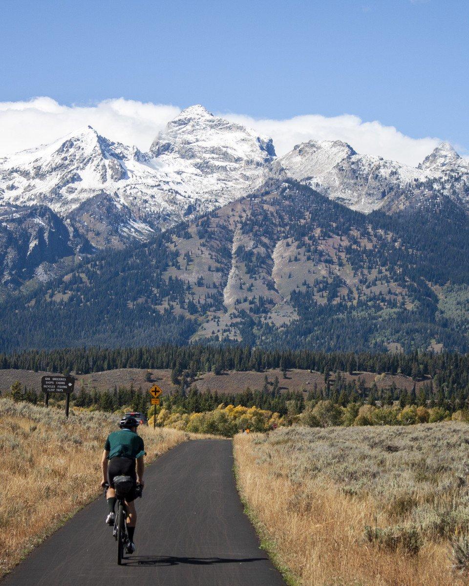 Cyclist in green jersey riding toward snow-capped mountains on remote road through golden grassland