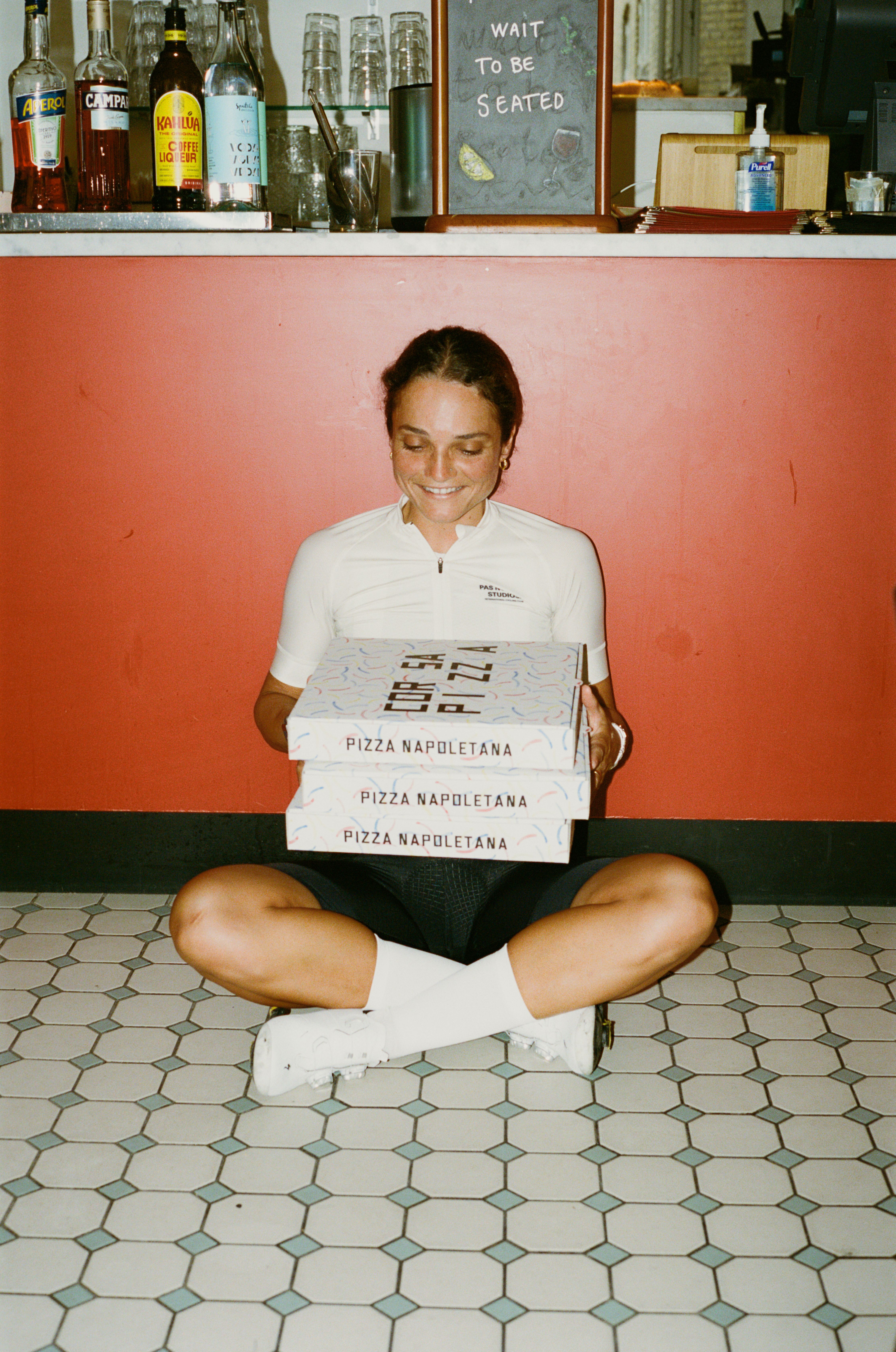 Model wearing white cycling polo with small chest branding, sitting cross-legged in restaurant holding pizza boxes