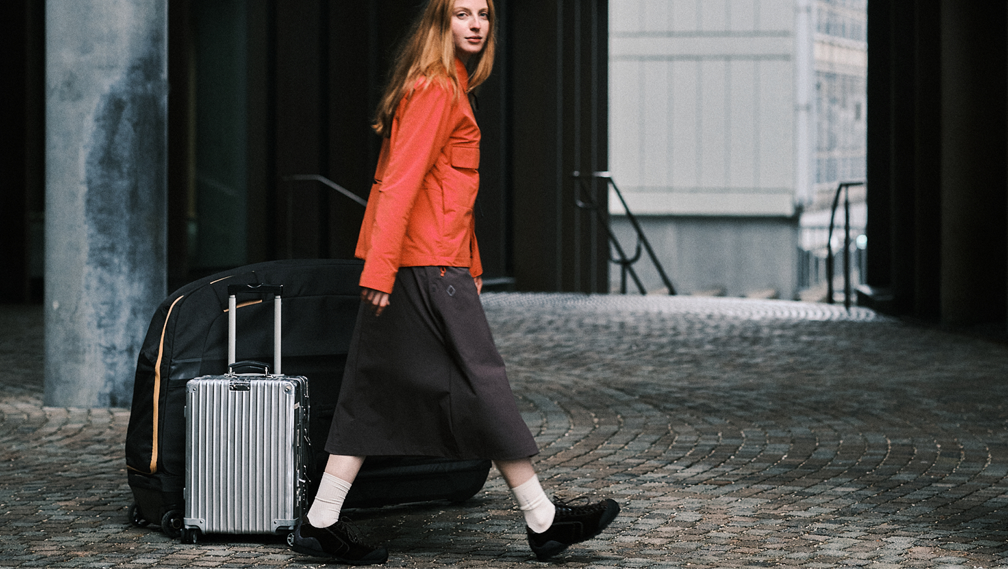 Model wearing orange jacket and dark pants walking with suitcase on cobblestone street