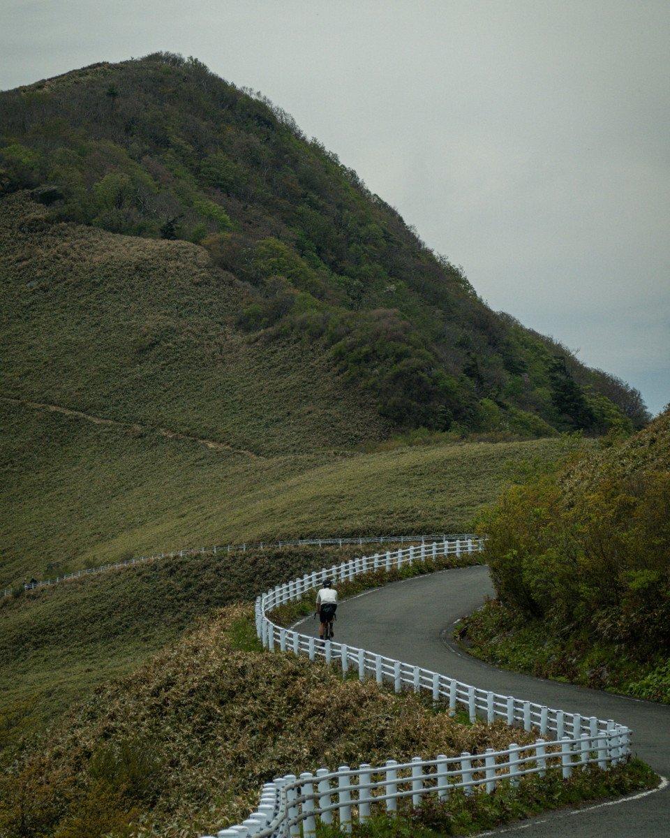 Cyclist on winding mountain road with white guardrails and green hills under overcast sky