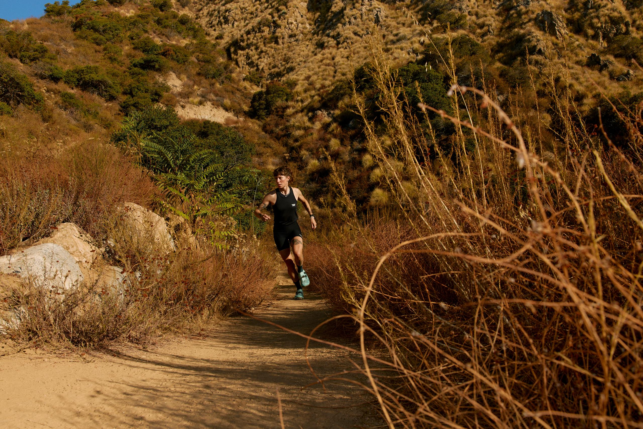Model wearing black sleeveless running top running on dirt trail through Mediterranean landscape