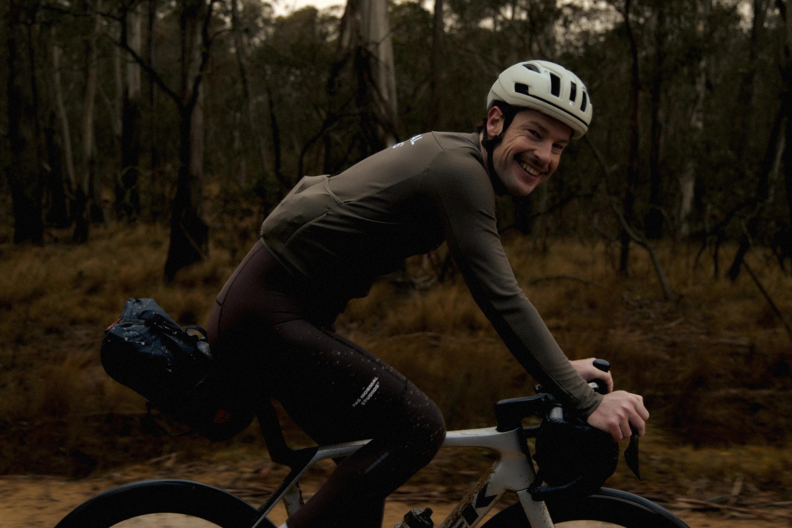 Model wearing brown long sleeve jersey and black tights while cycling through wooded terrain