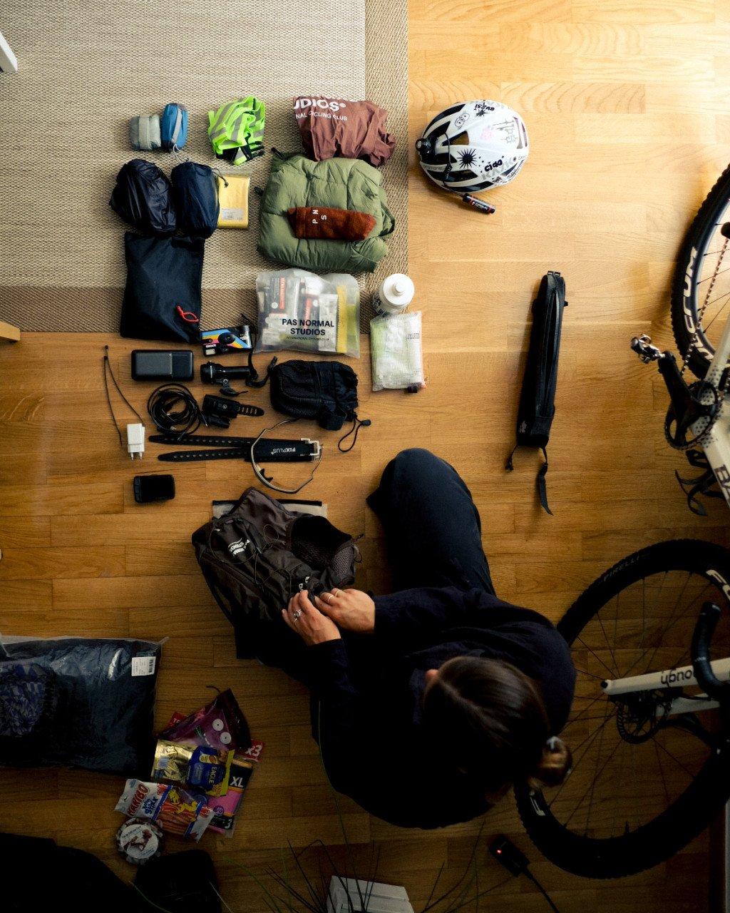 Cyclist organizing gear on wooden floor including packable jackets, helmet, and cycling equipment for bikepacking trip