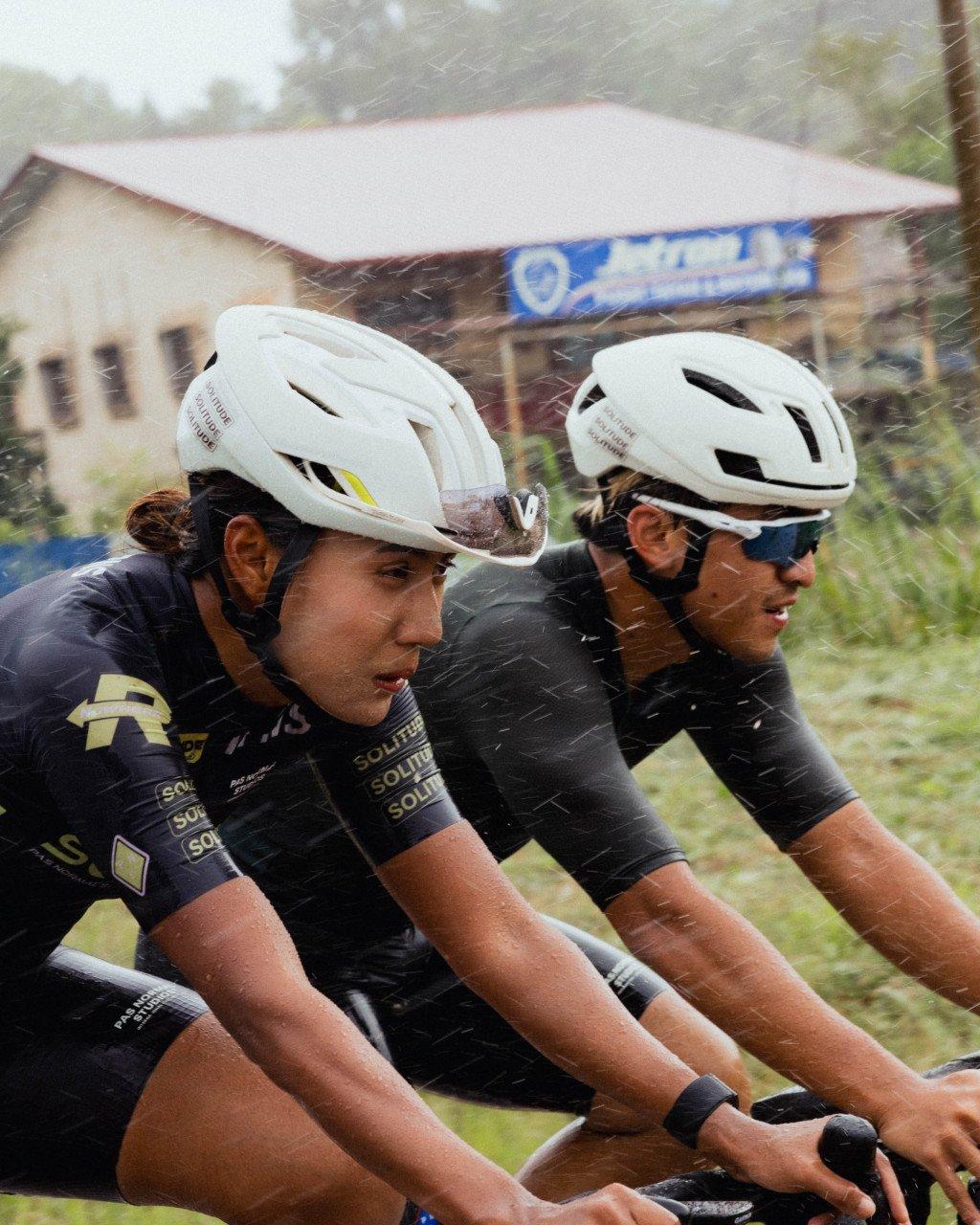 Two cyclists in dark racing jerseys riding in heavy rain with white helmets