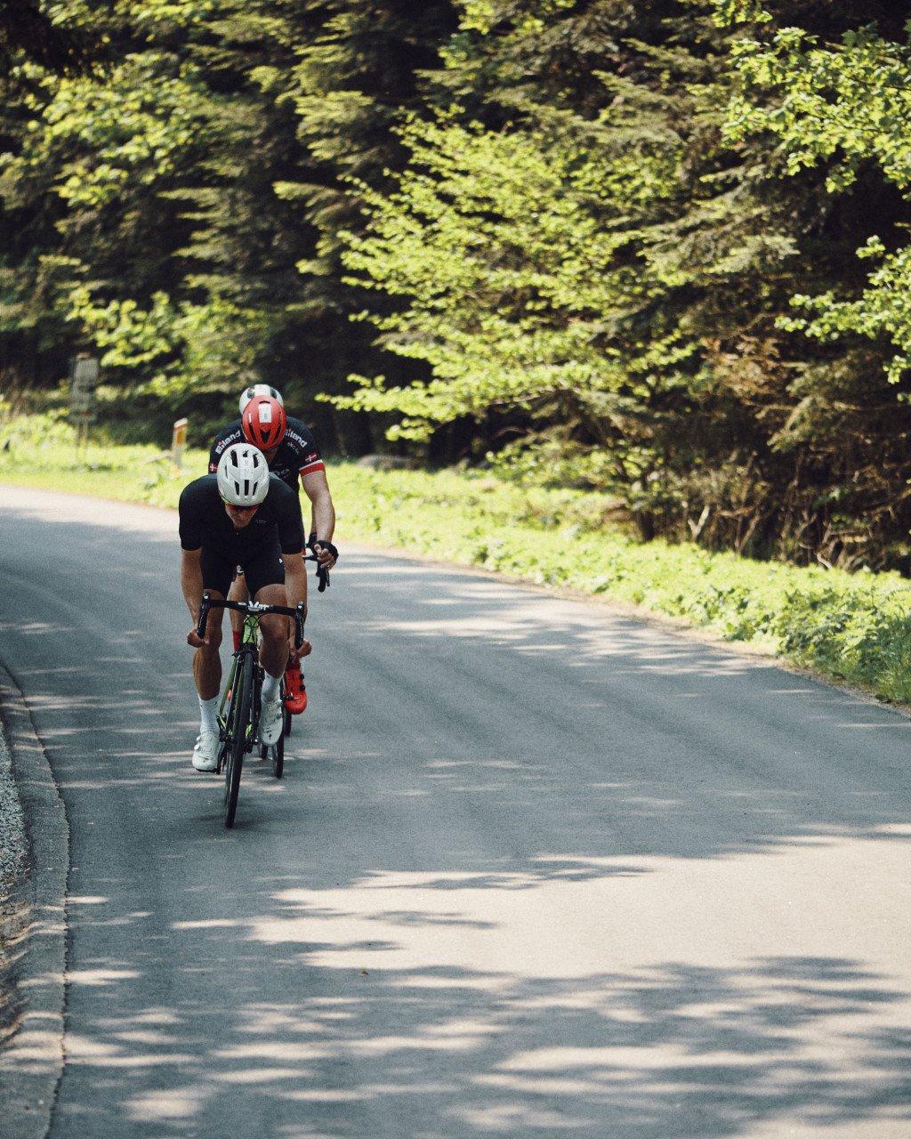Model wearing black cycling apparel in aerodynamic position on time trial bike on tree-lined road