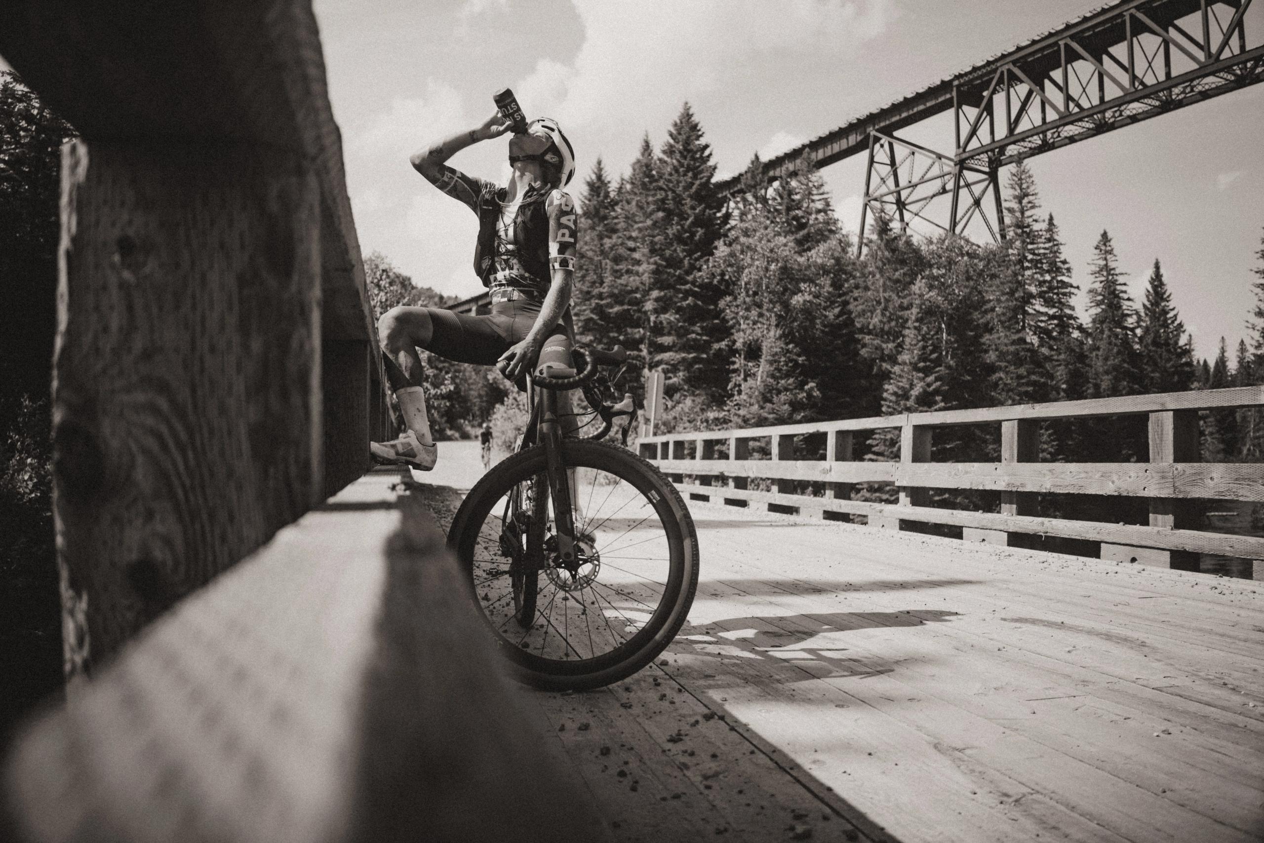 Model drinking from bottle while cycling across bridge in black and white outdoor setting