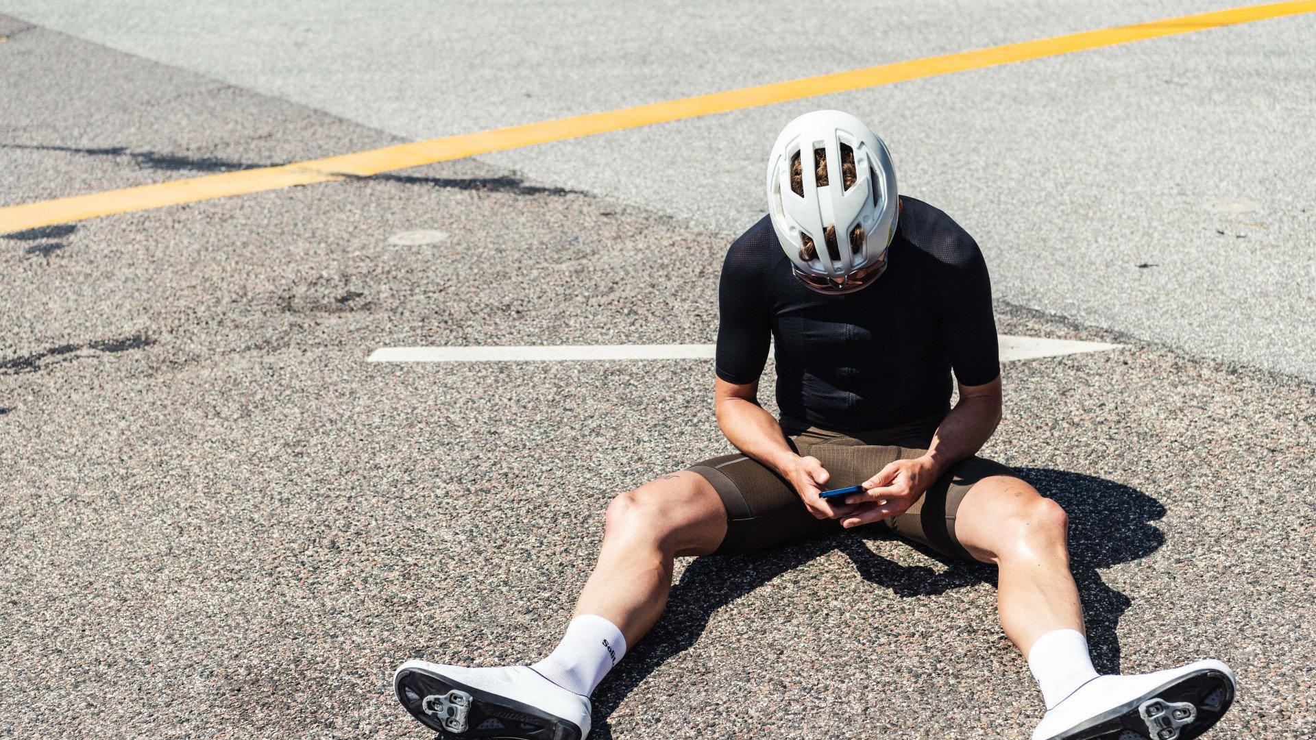 Model wearing black cycling jersey and olive shorts sitting on road with white helmet, holding phone