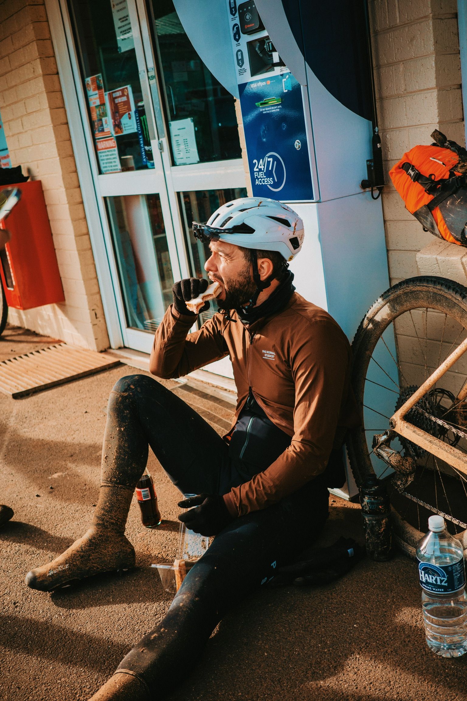 Model wearing brown long sleeve jersey and black cycling tights resting at fuel station with bicycle