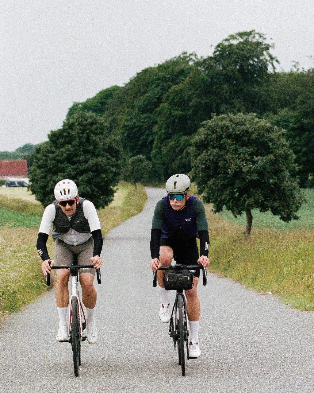 Two cyclists in aerodynamic position on time trial bikes wearing matching white helmets on rural road