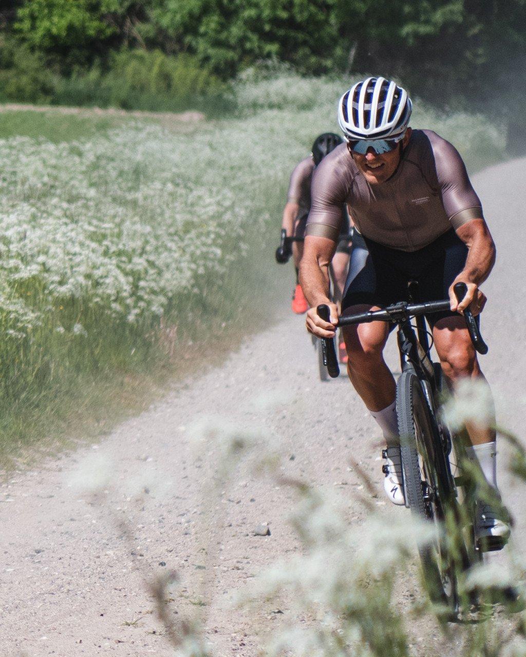 Model wearing brown jersey and black shorts cycling on gravel path through countryside