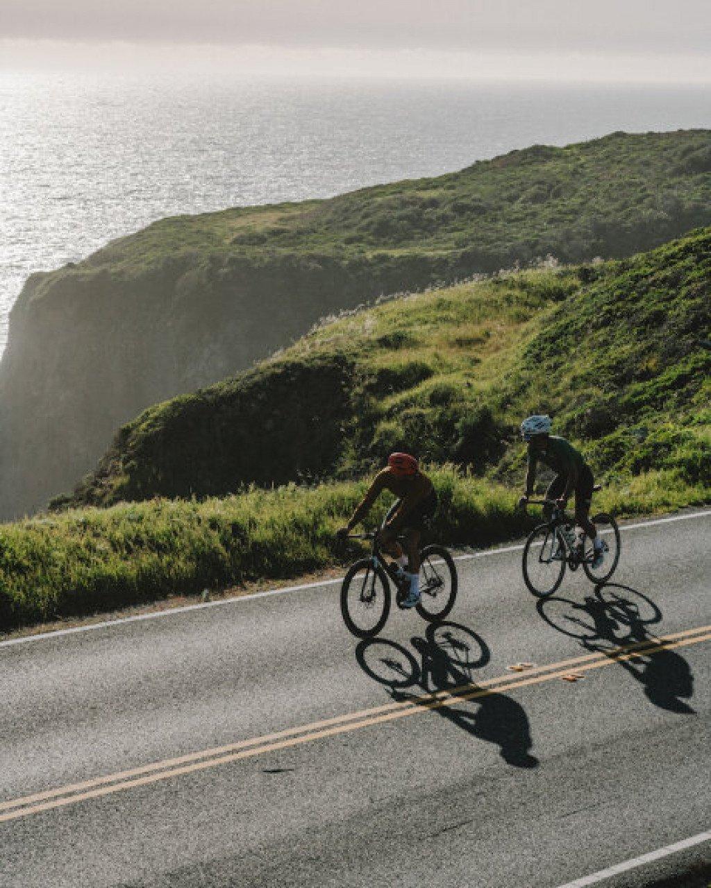 Two cyclists riding along coastal road with green hills and ocean backdrop in Sonoma, California