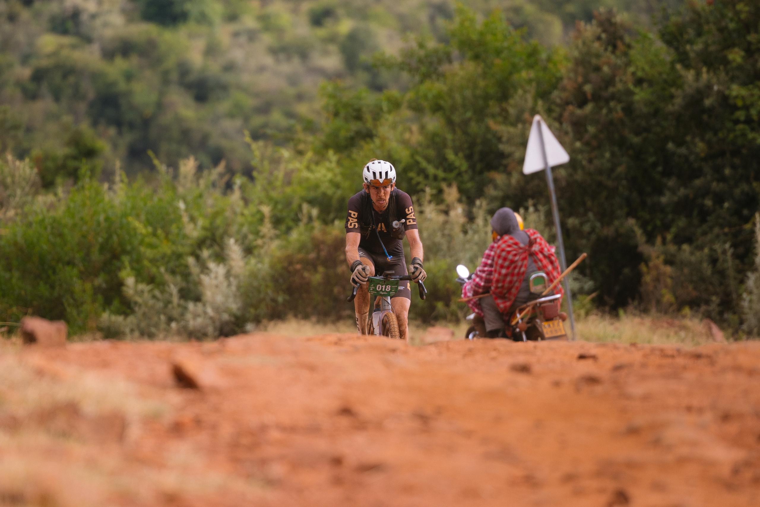 Cyclist in dark jersey racing gravel bike on dusty terrain with spectator in background
