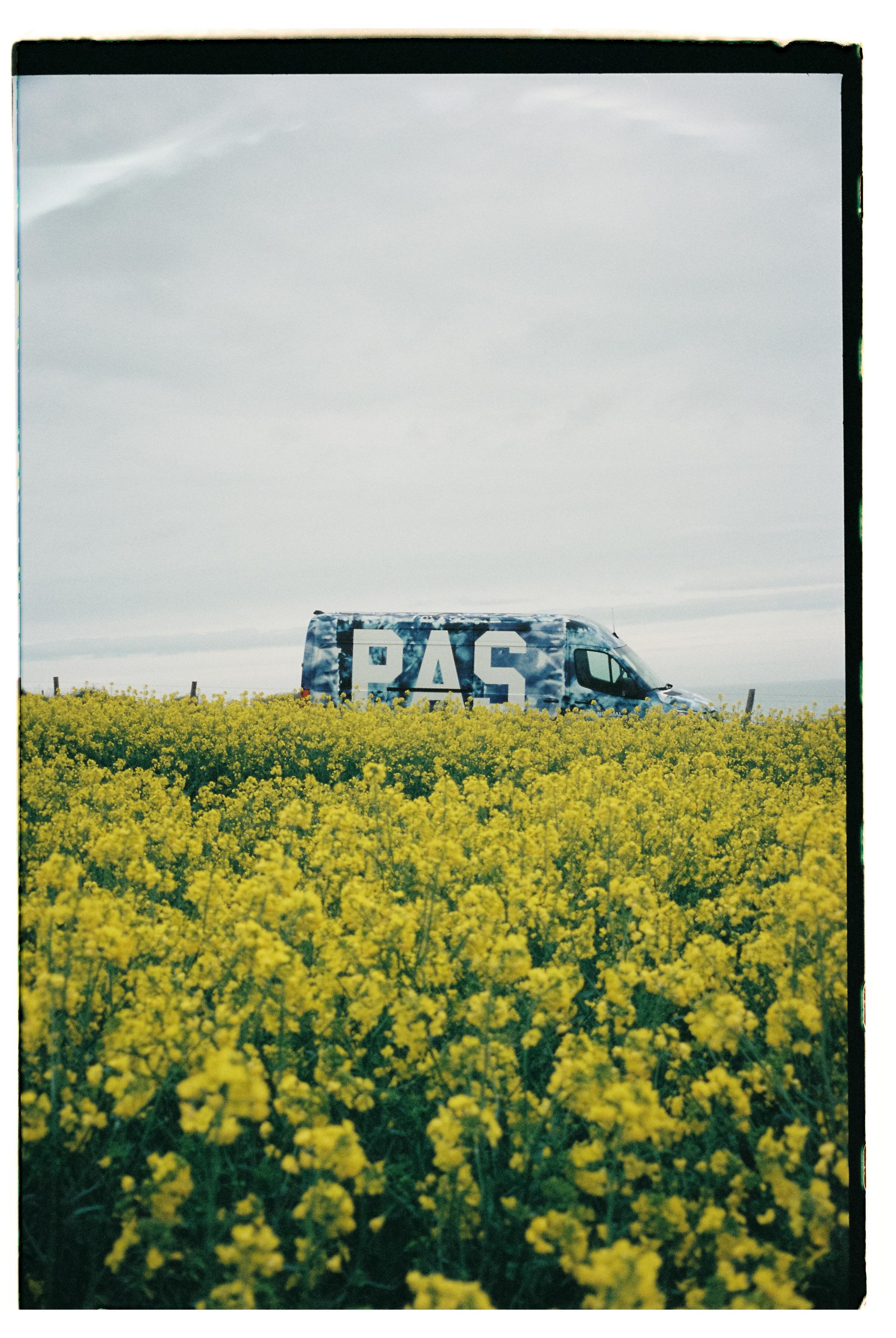 Blue camouflage van with logo in yellow wildflower field under overcast sky