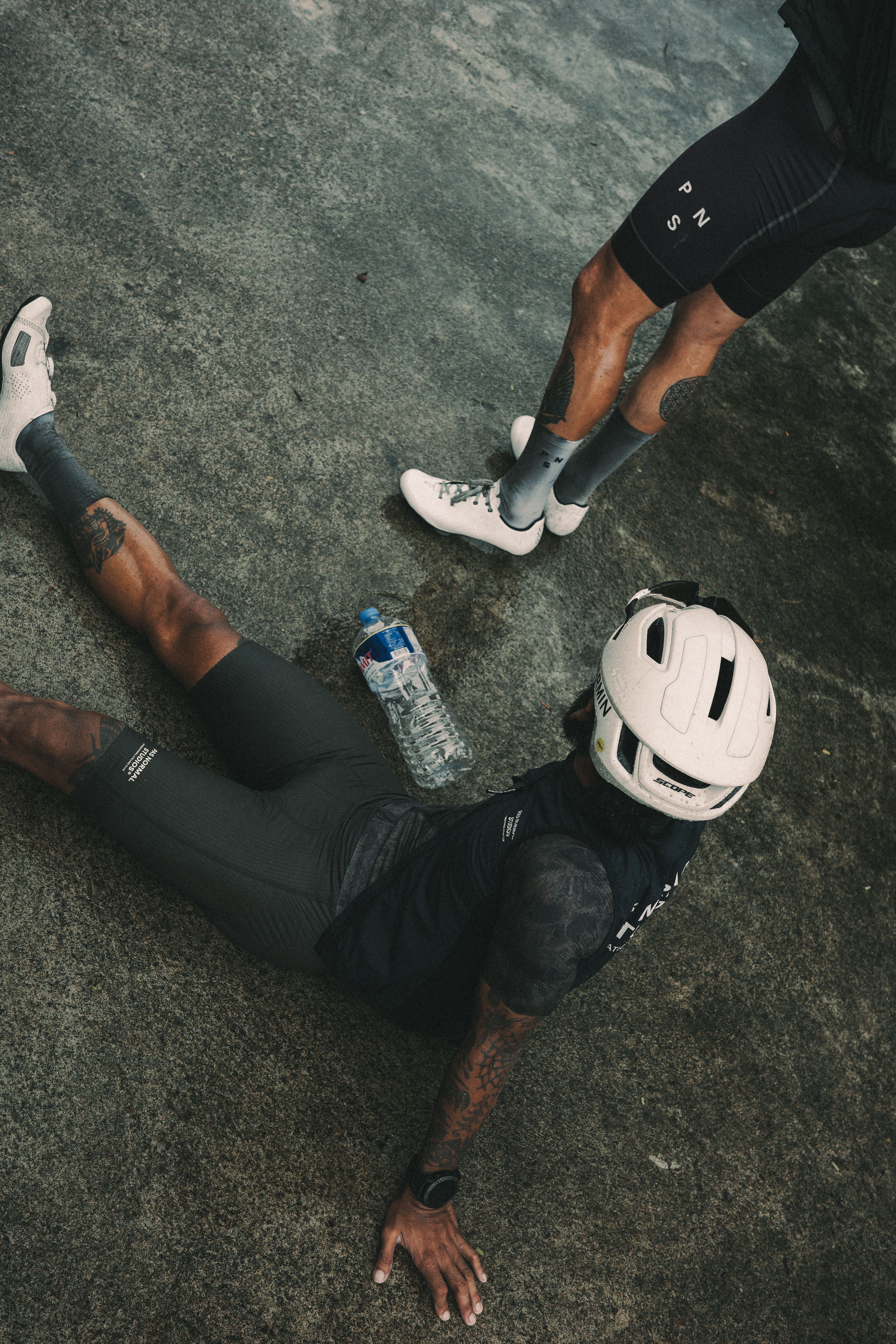 Cyclists in black cycling apparel resting on pavement with white helmet and water bottle