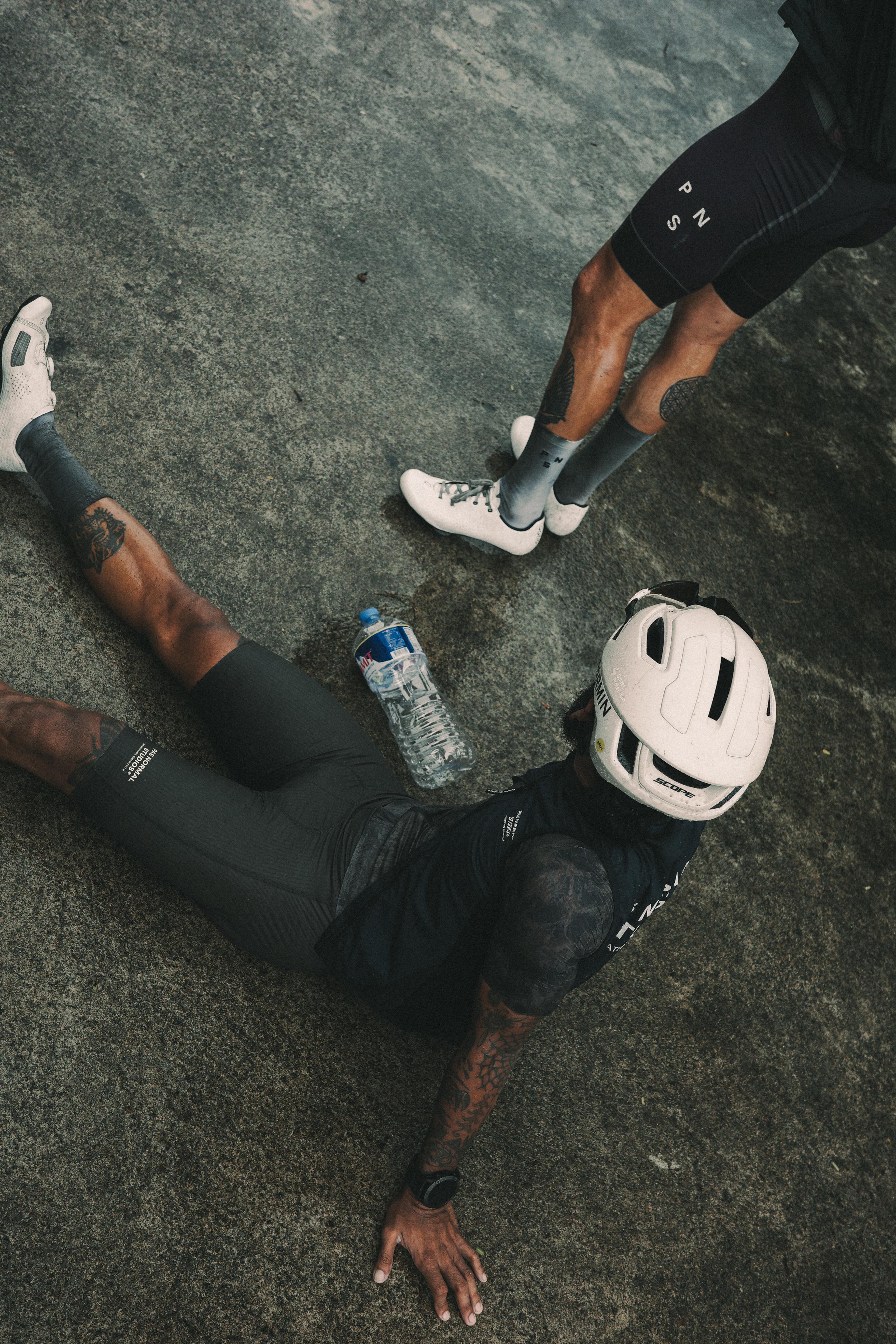 Cyclists in black cycling apparel resting on pavement with white helmet and water bottle