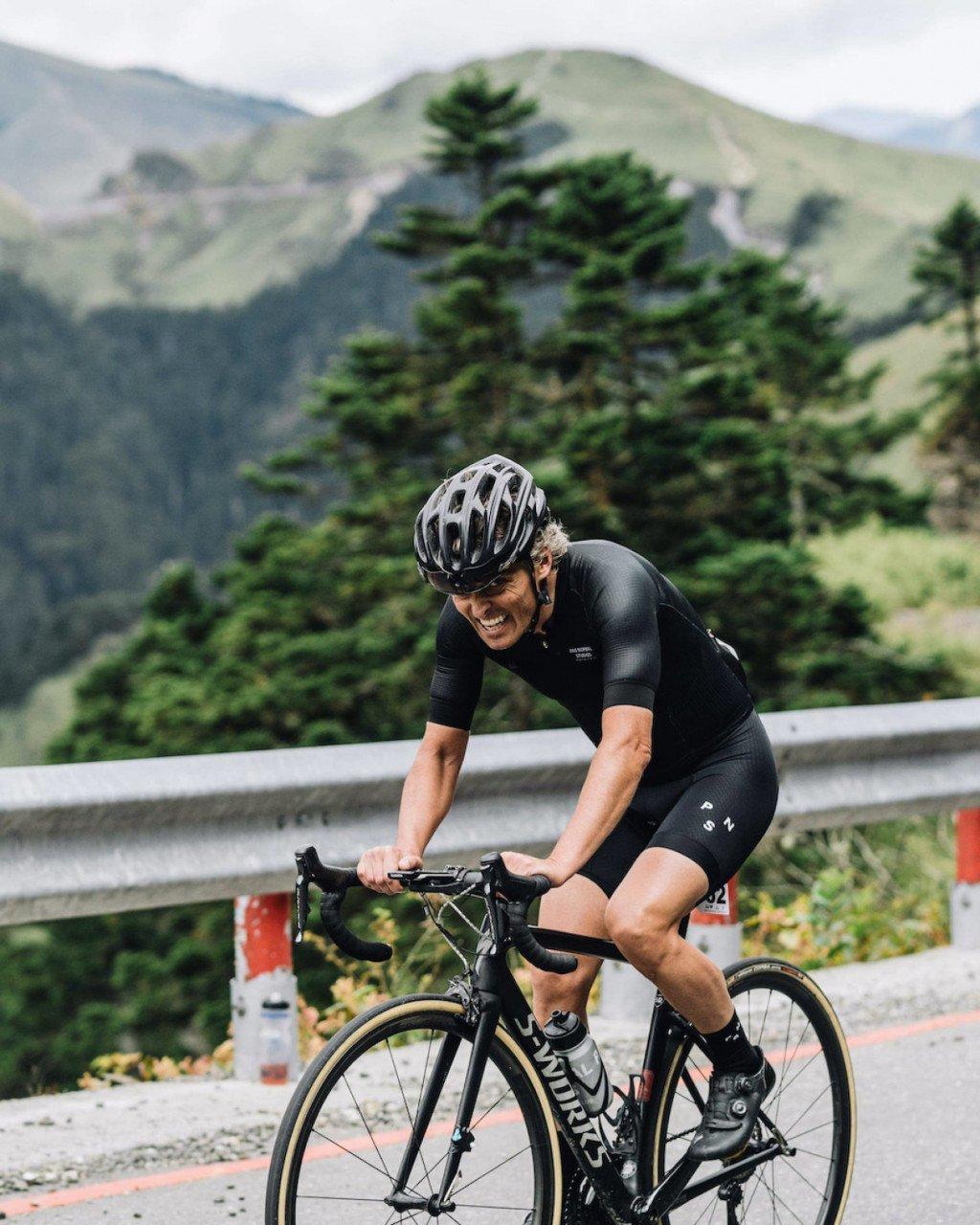 Model wearing black cycling kit in aerodynamic position on road bike in mountainous landscape