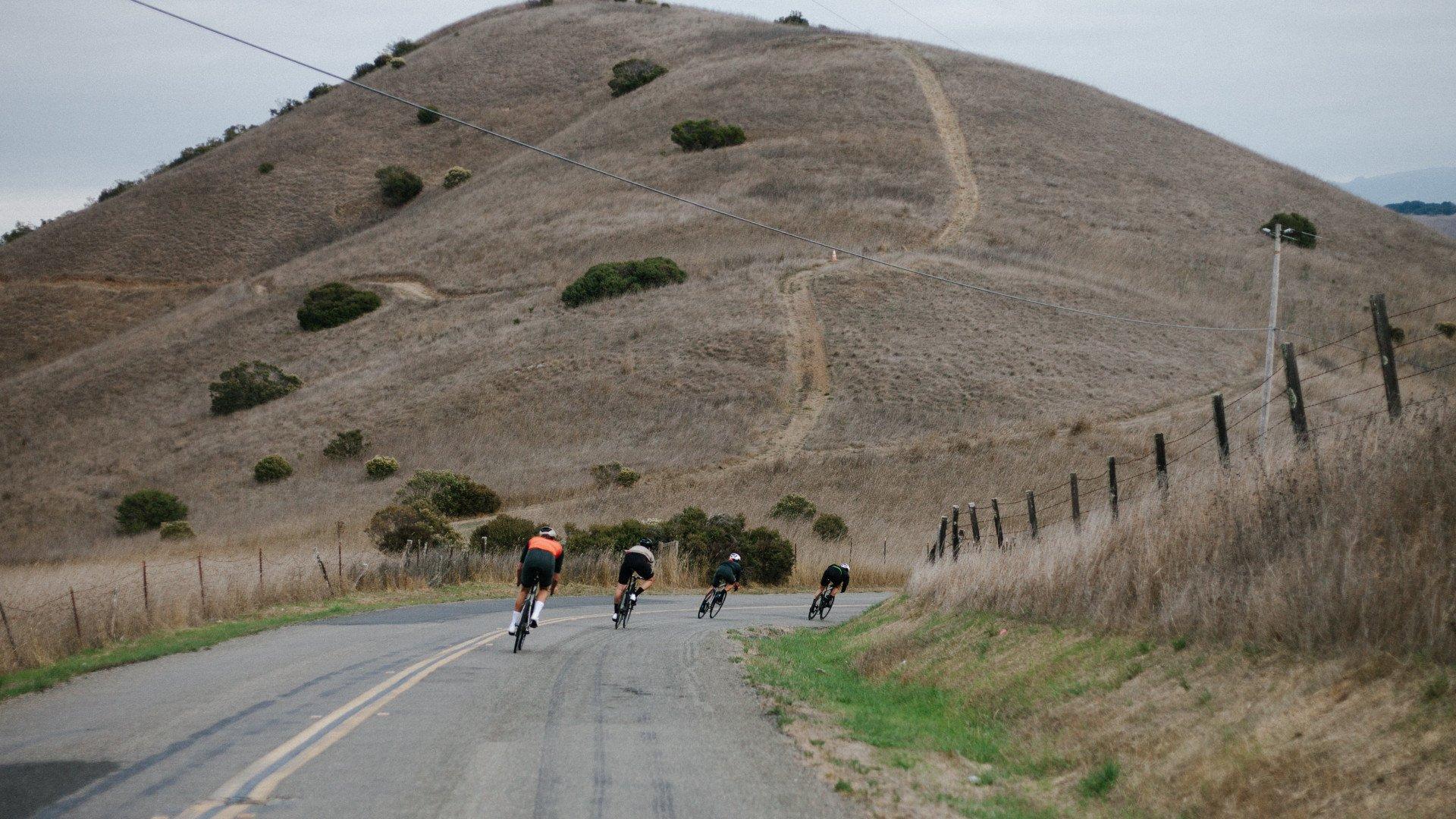Four cyclists in aerodynamic position riding on rural road with rolling hills in background