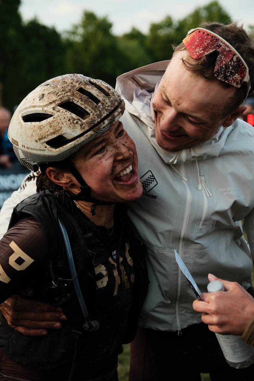 Two muddy cyclists celebrating after gravel race, one wearing helmet and dark jersey, other in light windbreaker