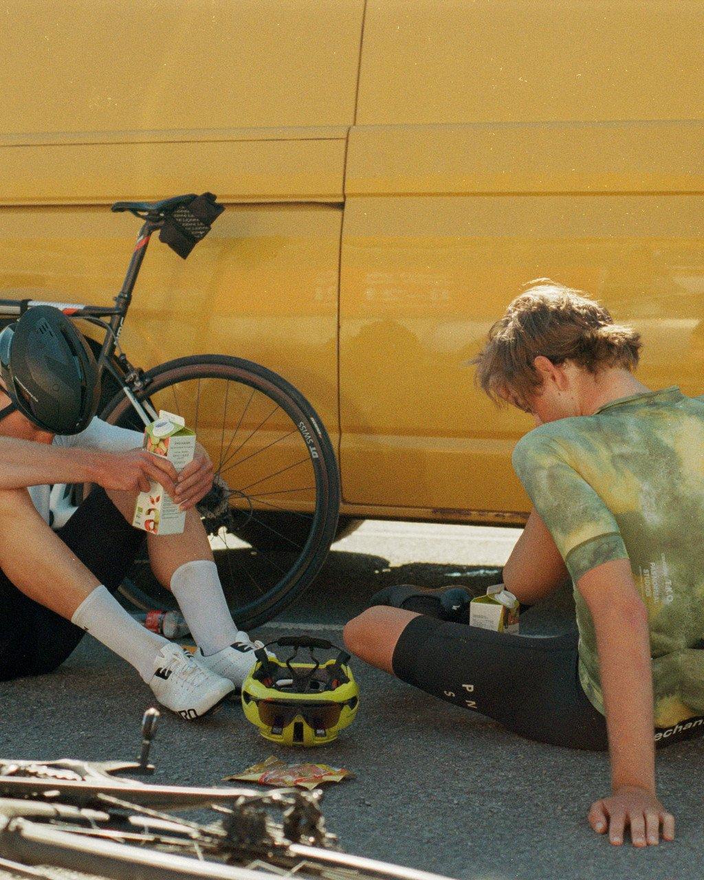 Female cyclists resting with bikes, one wearing tie-dye jersey, post-ride scene with gear