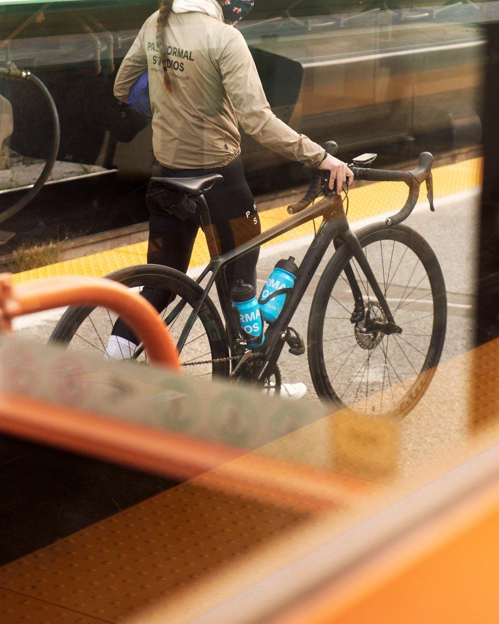 Model wearing beige jacket with back branding and black cycling tights with bike at train station platform