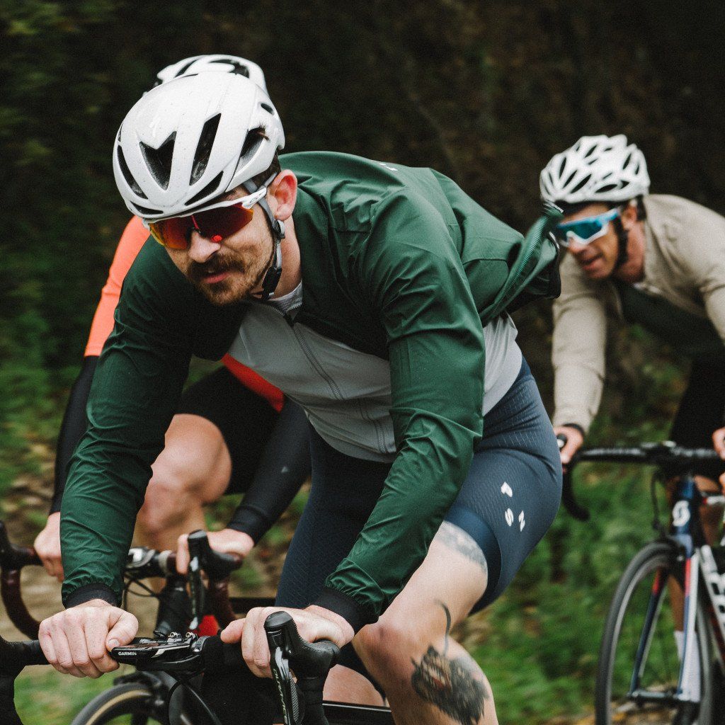 Model wearing green cycling jacket and navy bib shorts with white helmet riding road bike outdoors