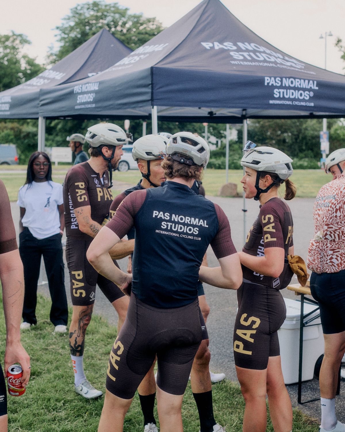 Cyclists in brown and navy team kit with helmets gathered under branded tent at cycling event