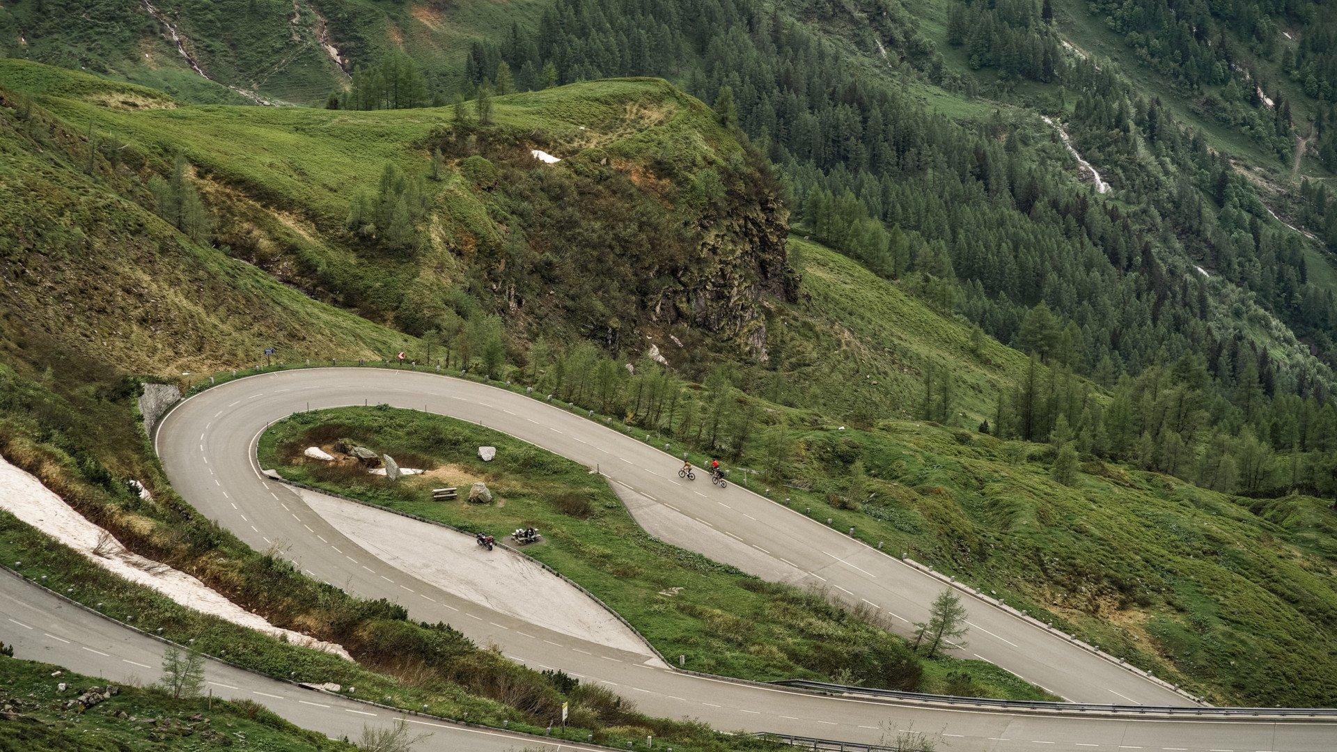 Cyclists on winding mountain hairpin turn surrounded by green hills and forests