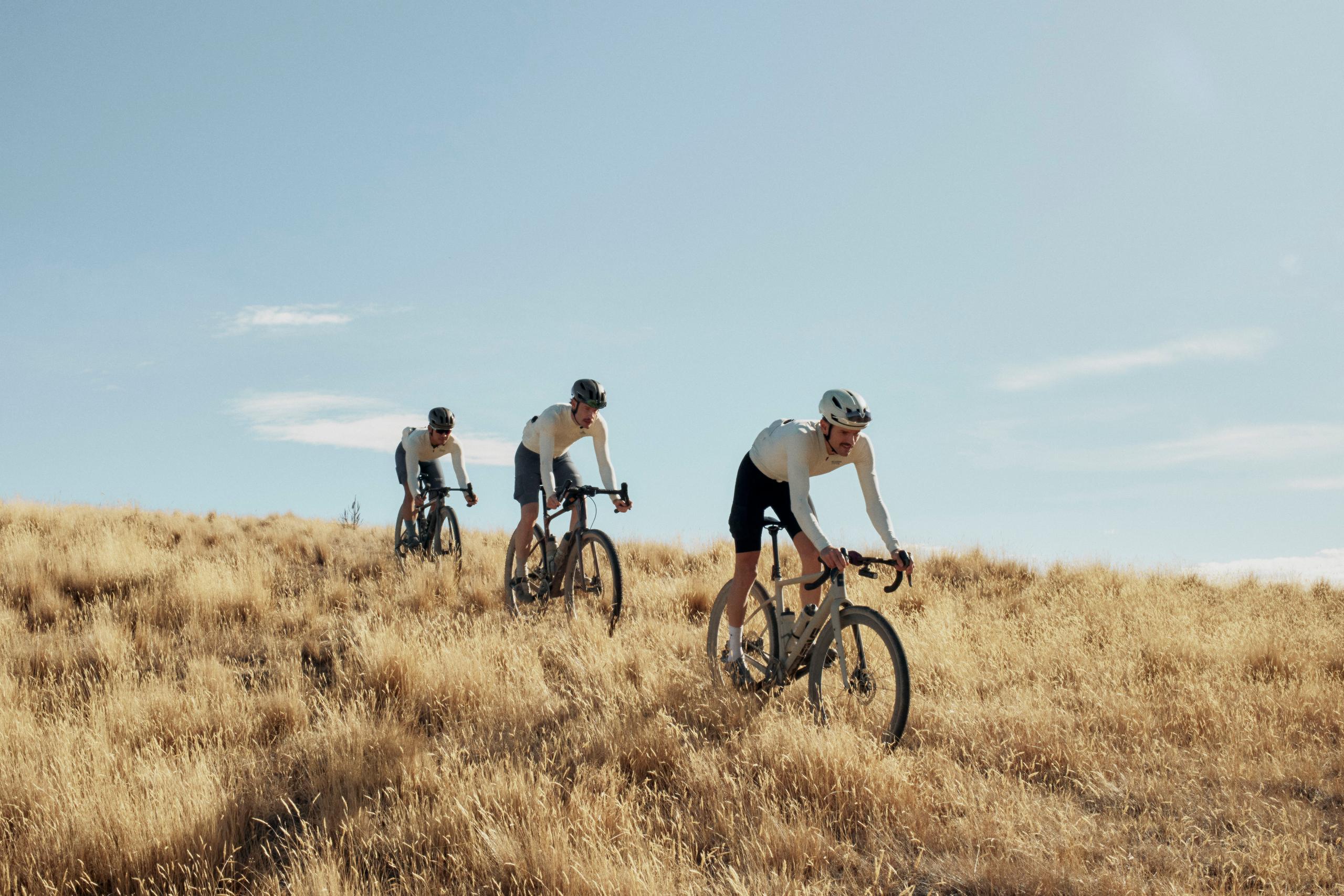 Three cyclists in aerodynamic position riding through golden grassland under blue sky