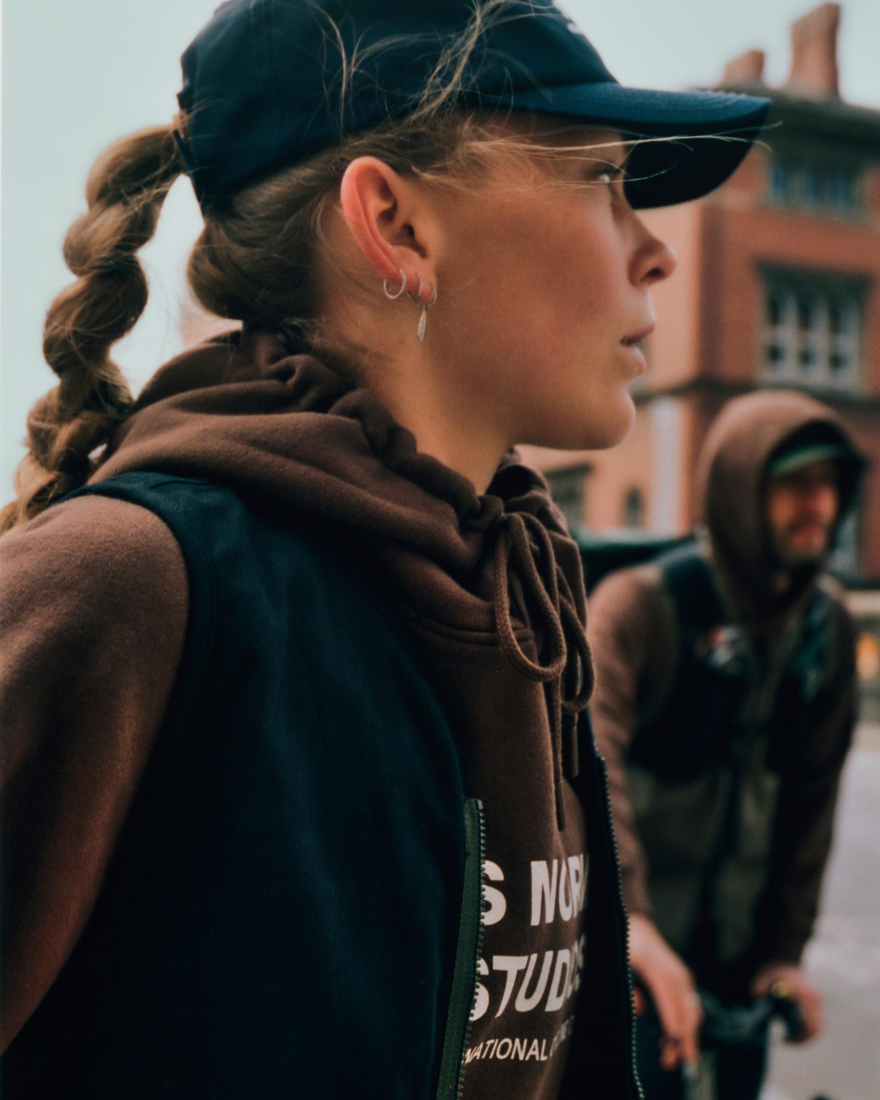 Model wearing brown hoodie with chest branding and black cap in urban setting