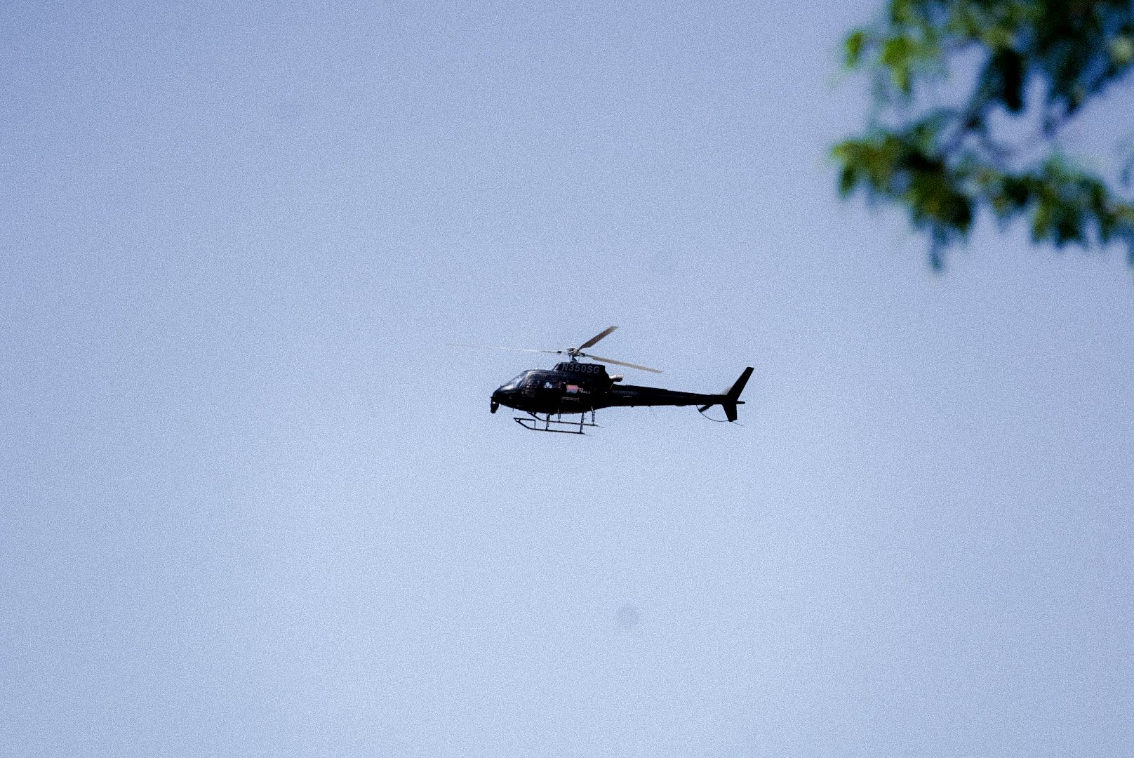 Black helicopter flying against blue sky with trees visible in corner