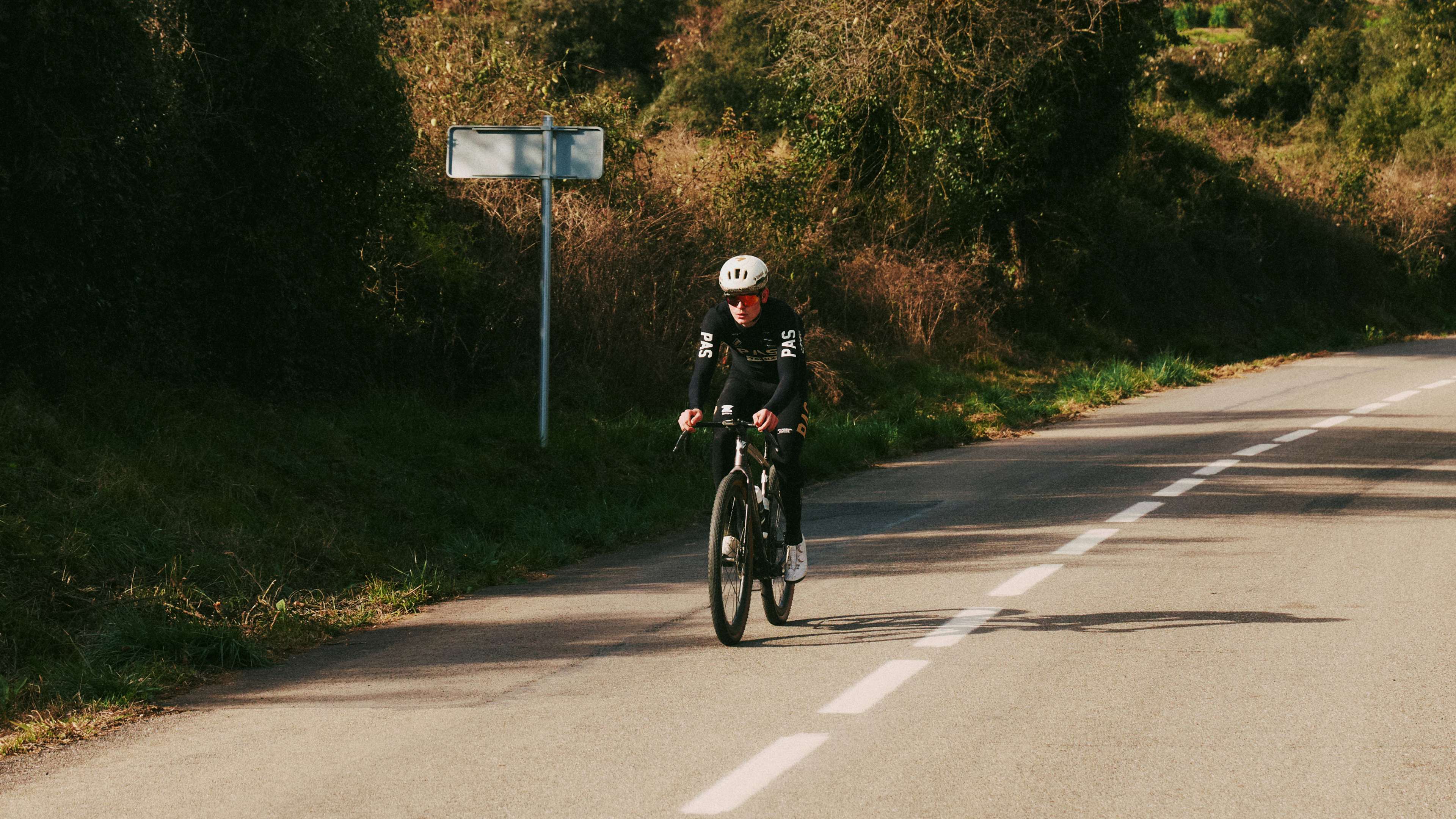 Model wearing black cycling outfit with branding on aerodynamic bike during time trial on rural road
