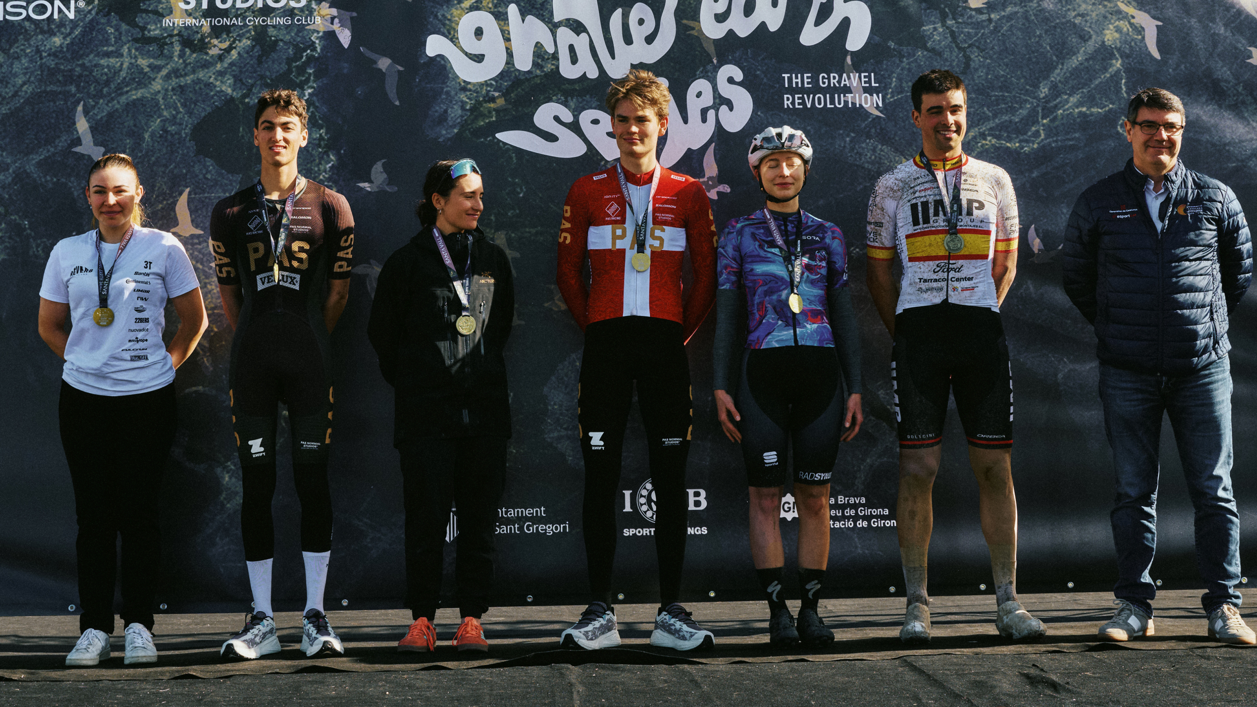 Cyclists on podium wearing various team jerseys with medals at gravel racing event