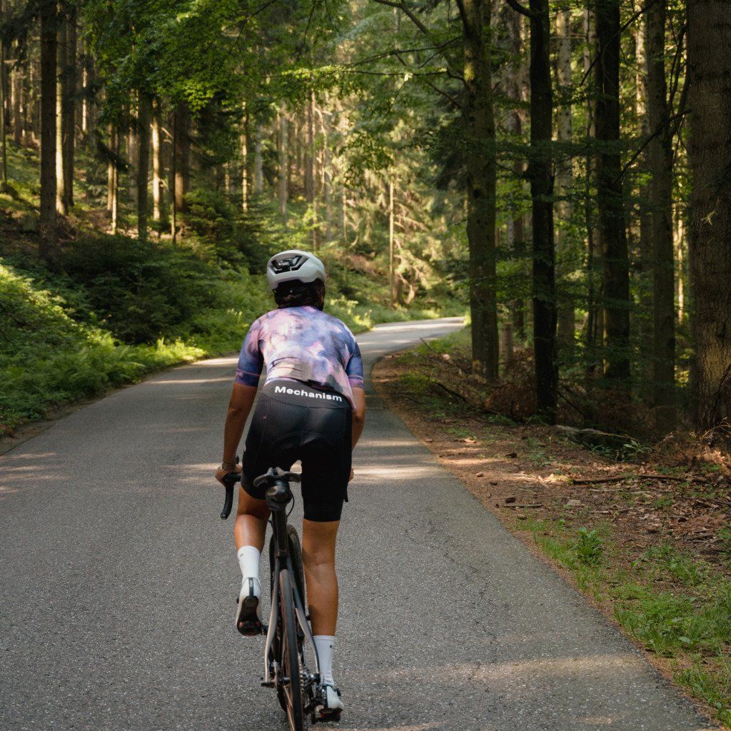 Model wearing Mechanism jersey with tie-dye pattern and black shorts cycling through forest road